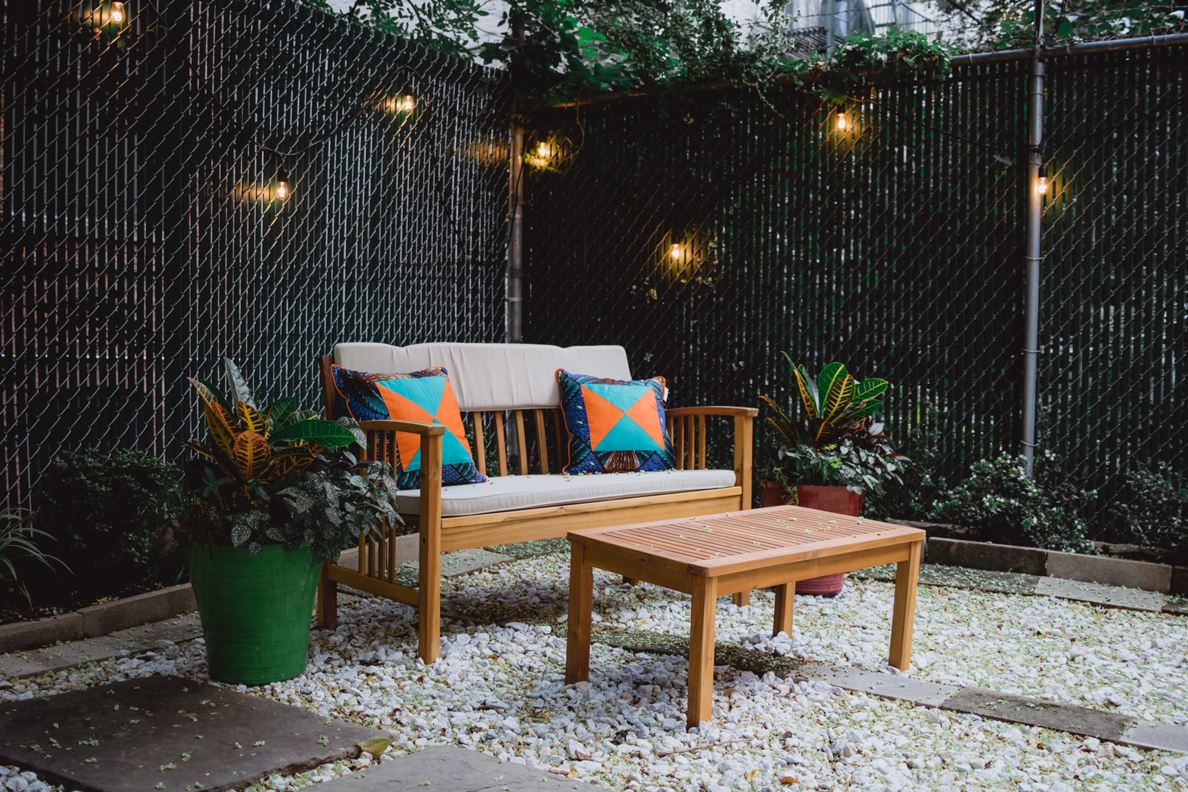A wooden sofa with colorful cushions and a matching coffee table sits in a gravel-covered patio surrounded by potted plants and a chain-link fence.