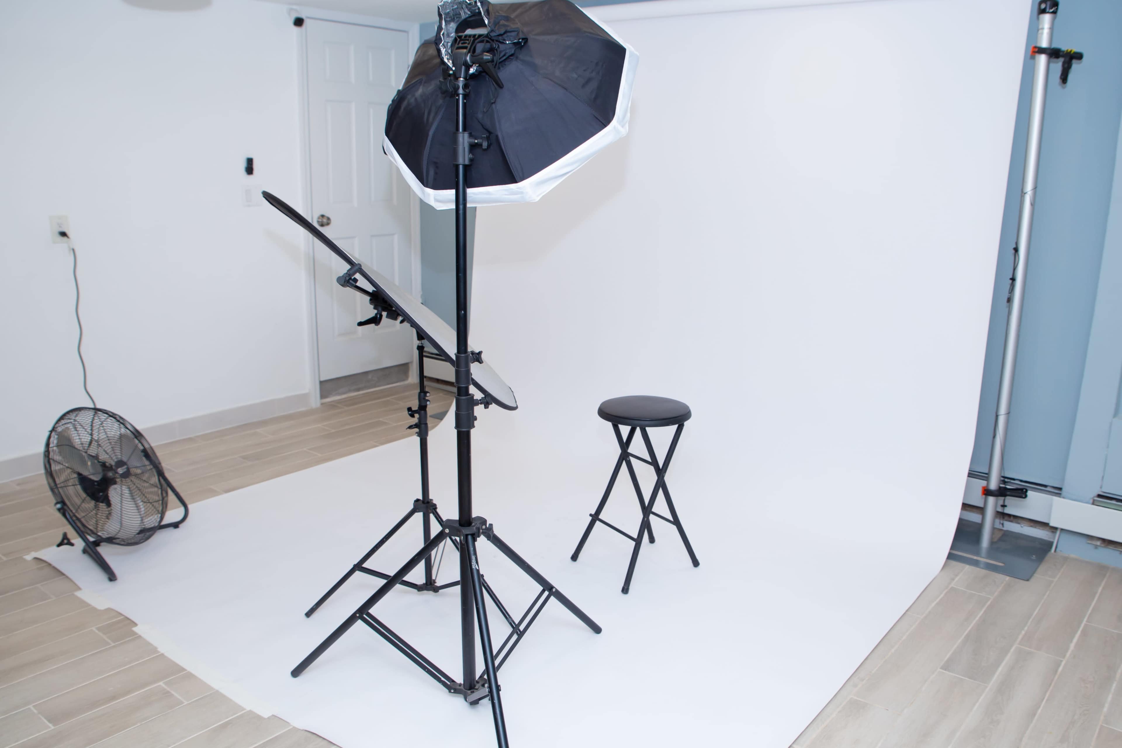 The image shows a photography studio setup featuring a black umbrella light, a dark stool, and a fan beside a backdrop of white paper.