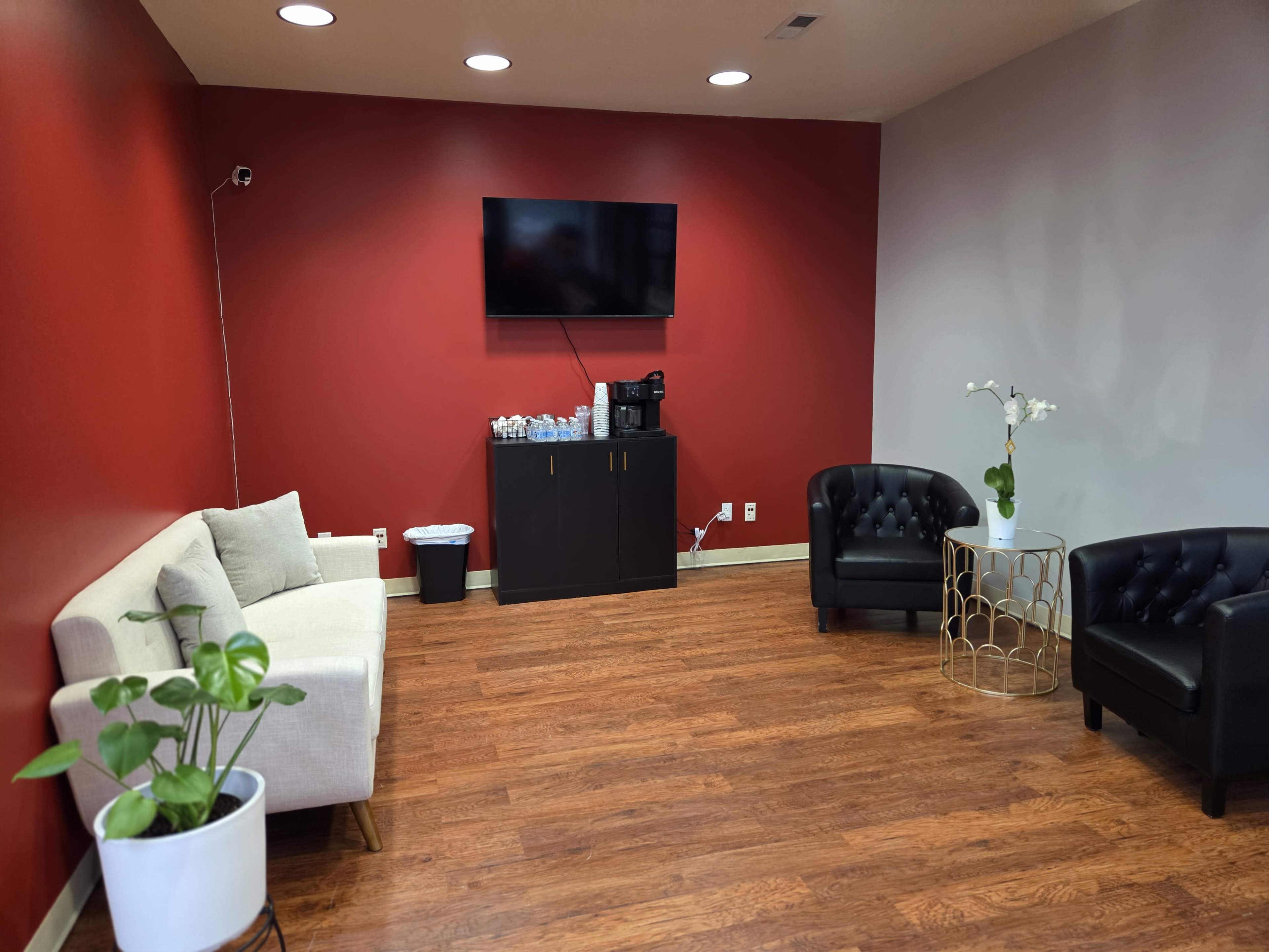The image shows a waiting room with a red accent wall, a beige sofa, two black chairs, a small round table, and a television mounted on the wall.
