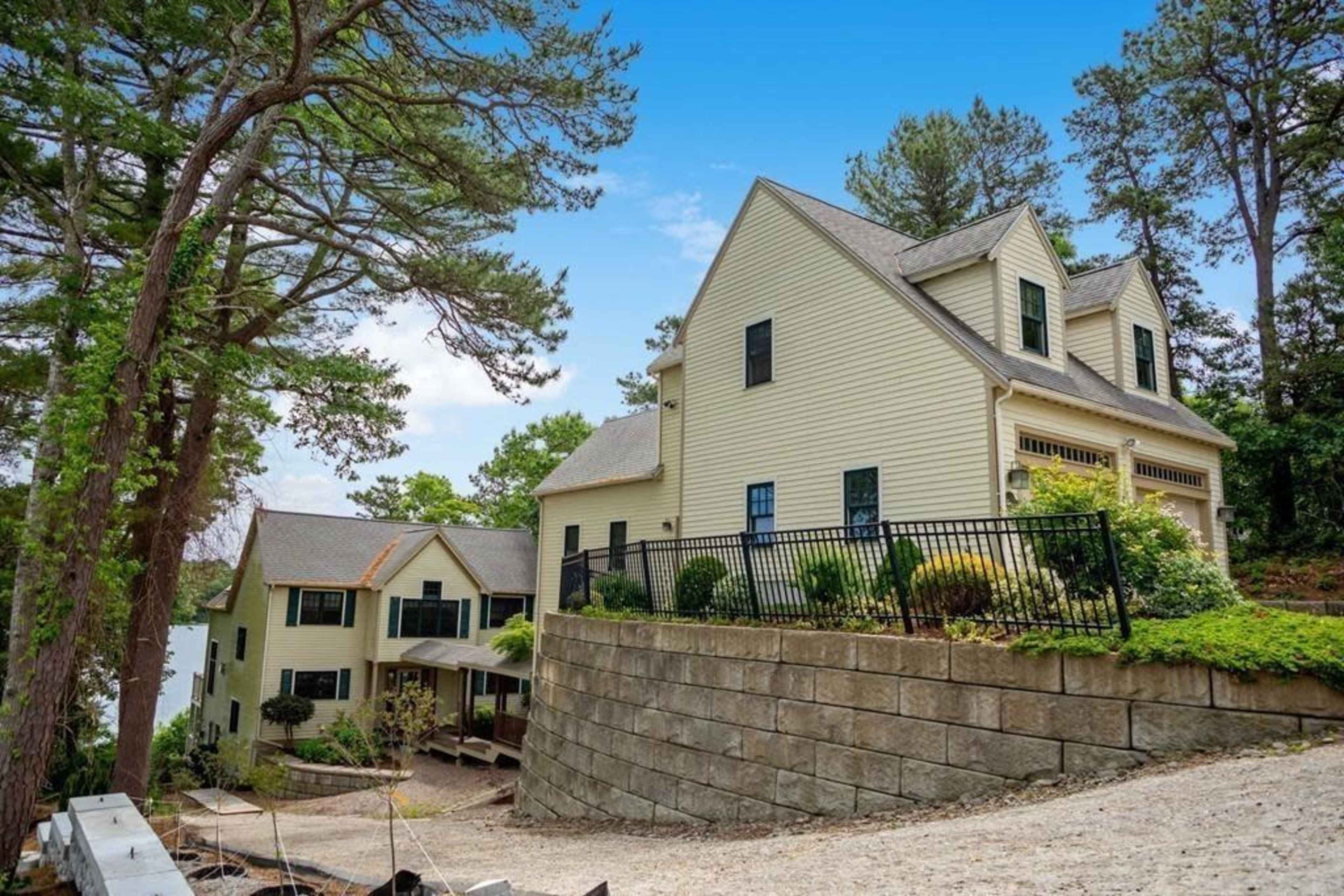 A two-story house with a light-colored exterior sits on a hillside, surrounded by trees and featuring a stone retaining wall.