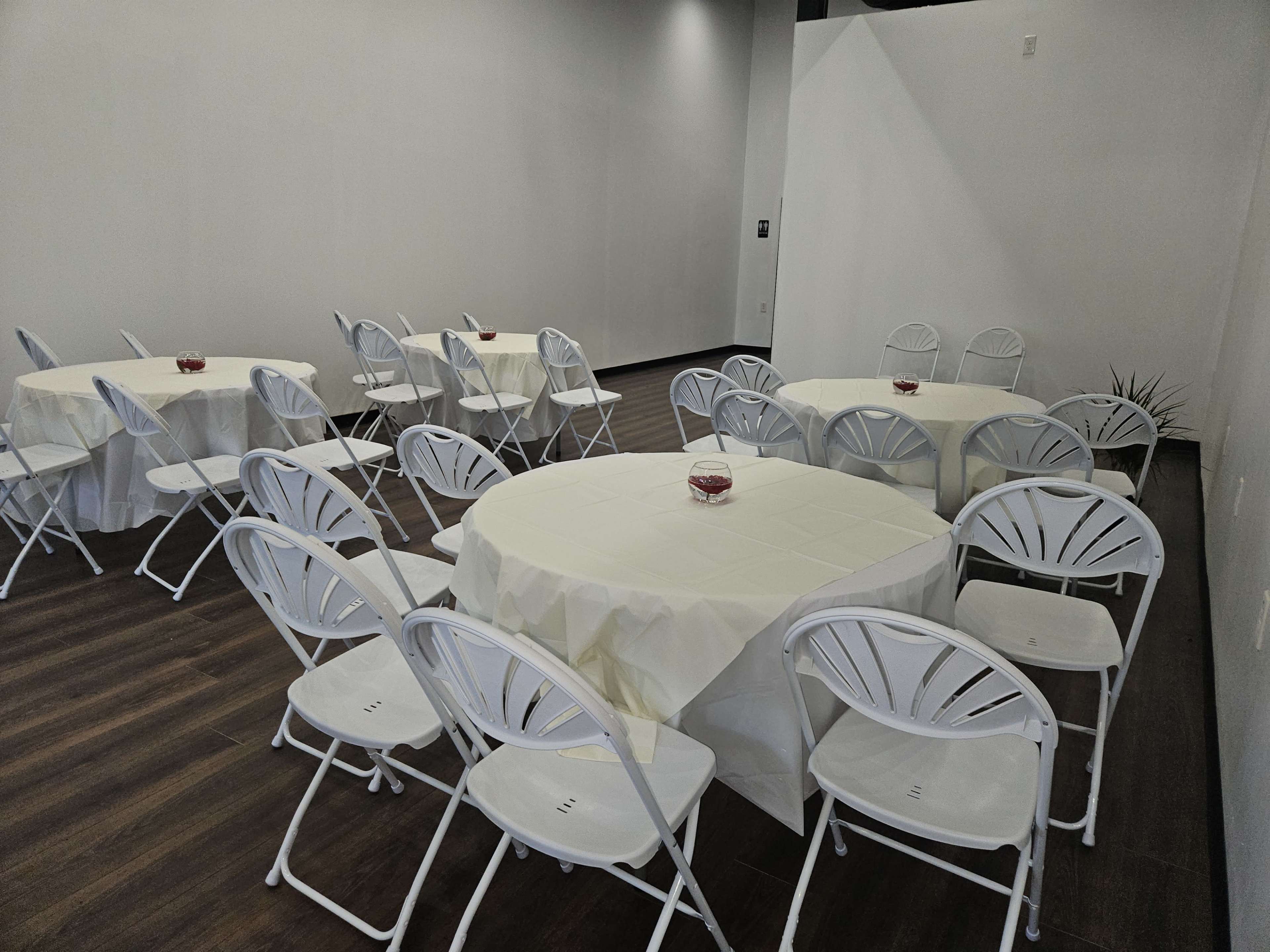 A room with several round tables covered with white tablecloths and surrounded by white folding chairs.