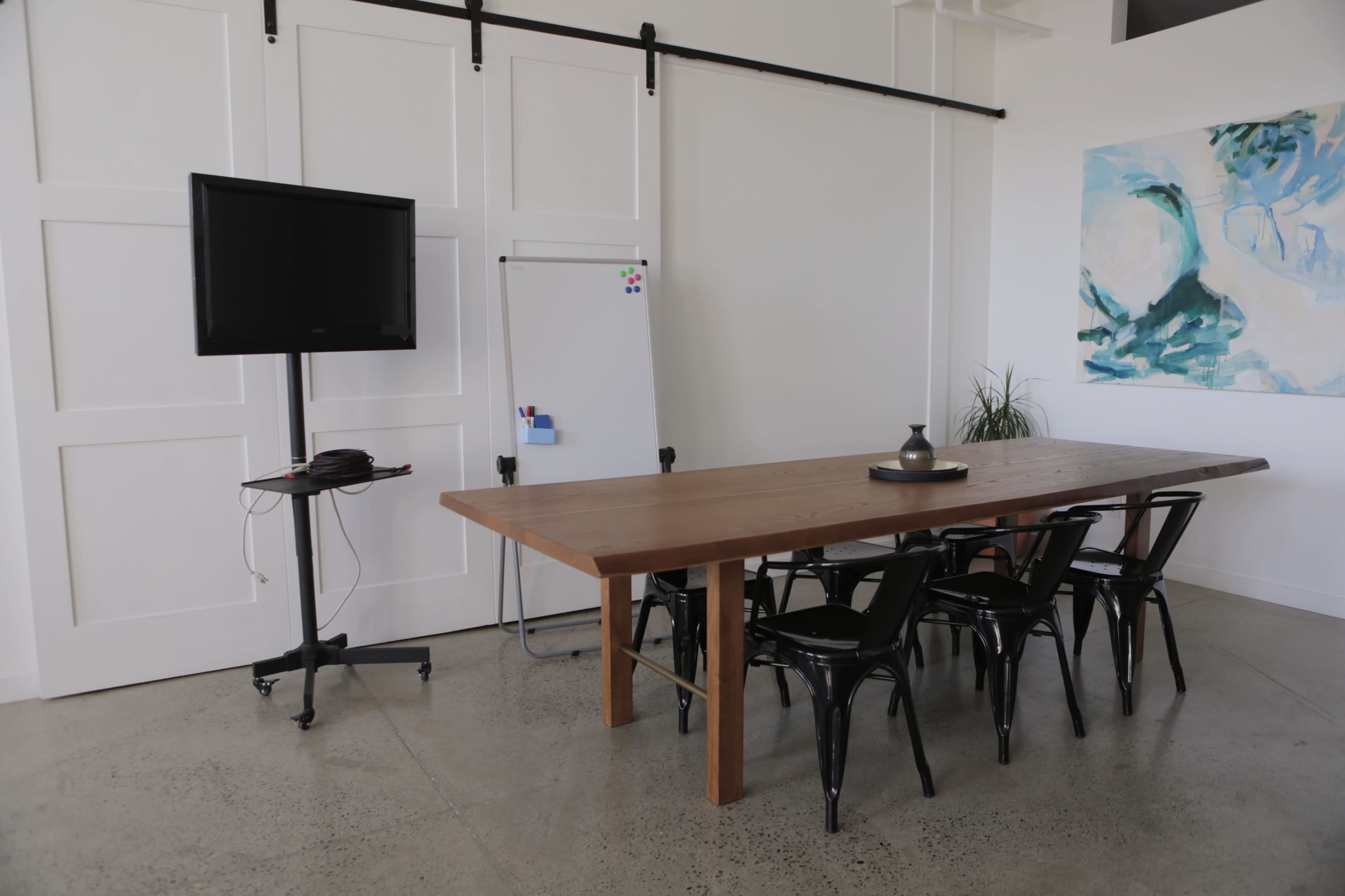 A conference room features a wooden table surrounded by black chairs, a television on a stand, a whiteboard, and a piece of wall art.