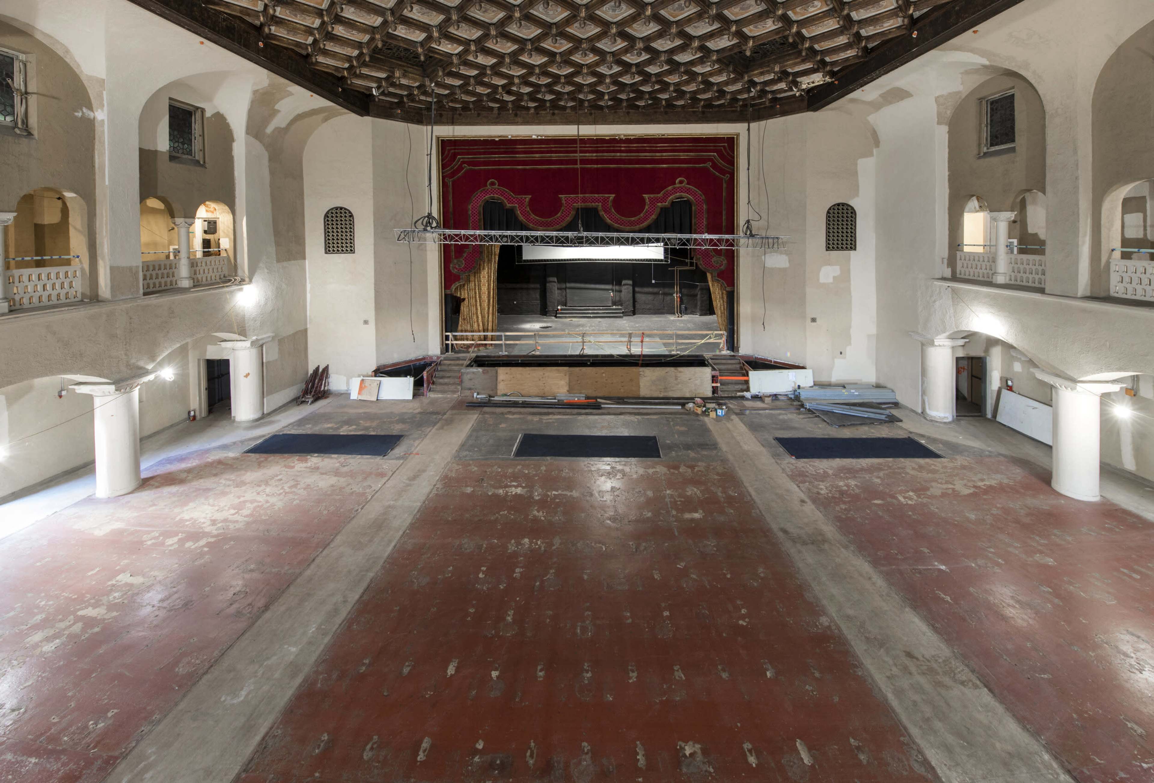 An empty theater with a stage at the front, adorned with red curtains, and a spacious, bare wooden floor.