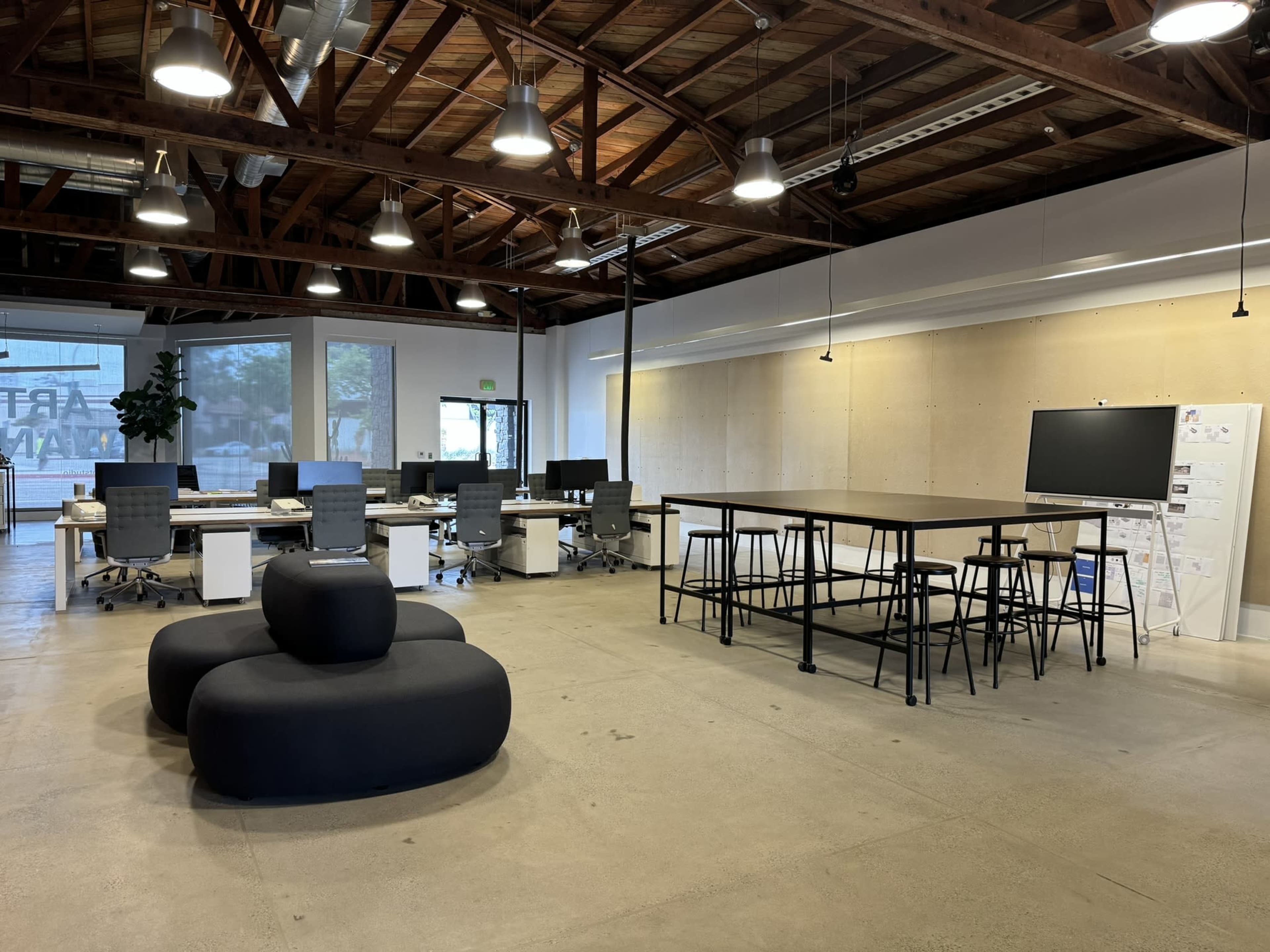 A modern office interior with desks, chairs, and a communal table under a wooden ceiling and bright lighting.