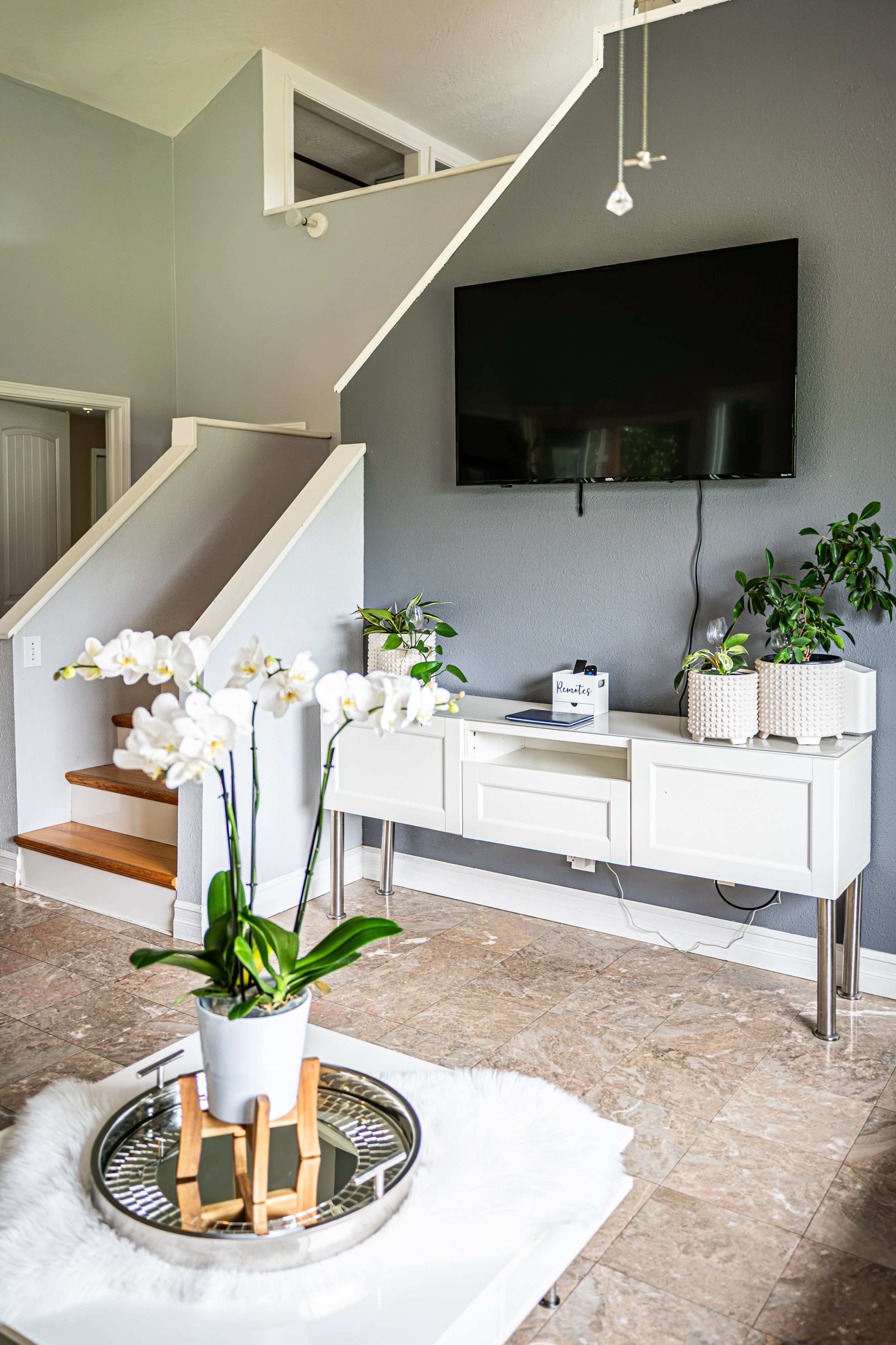 The image shows a modern living room with a white furniture set, a large television mounted on a gray wall, and a staircase in the background.