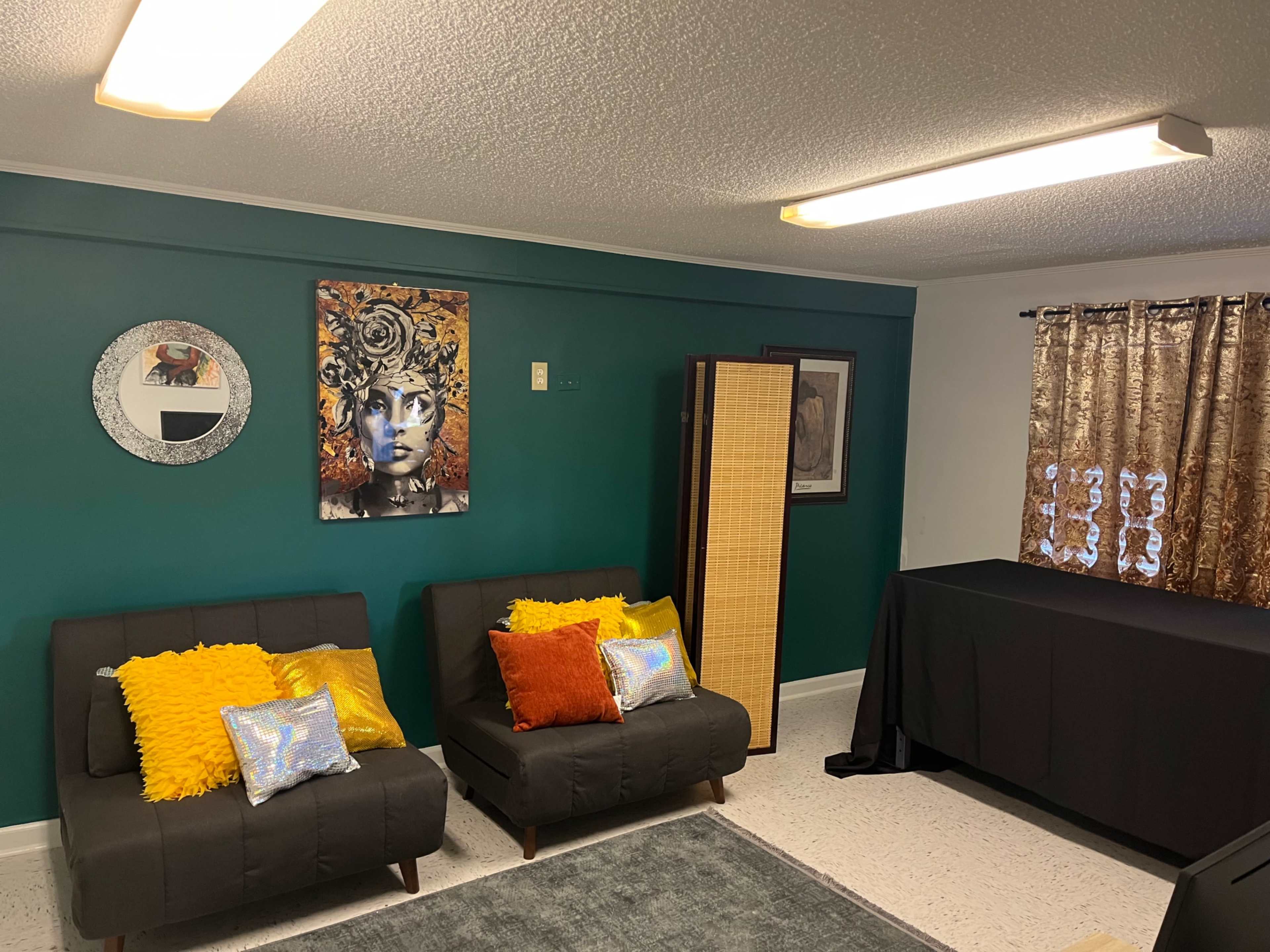 The room features two dark couches with colorful pillows, a decorative wall with artwork, and a folding screen beside a table under bright ceiling lights.
