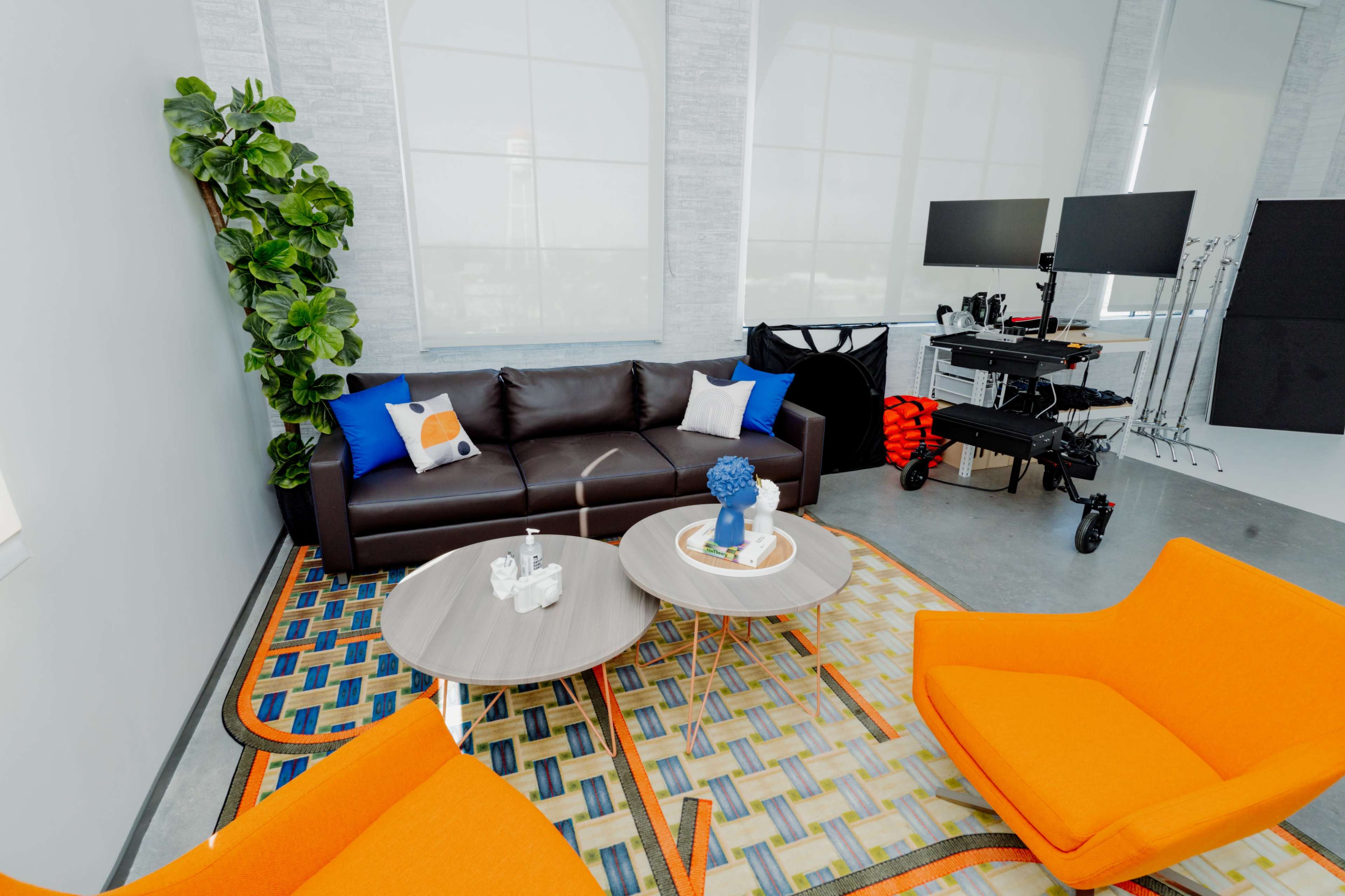A modern lounge area with a brown couch, two orange chairs, a circular coffee table, and a large window with light filtering through.