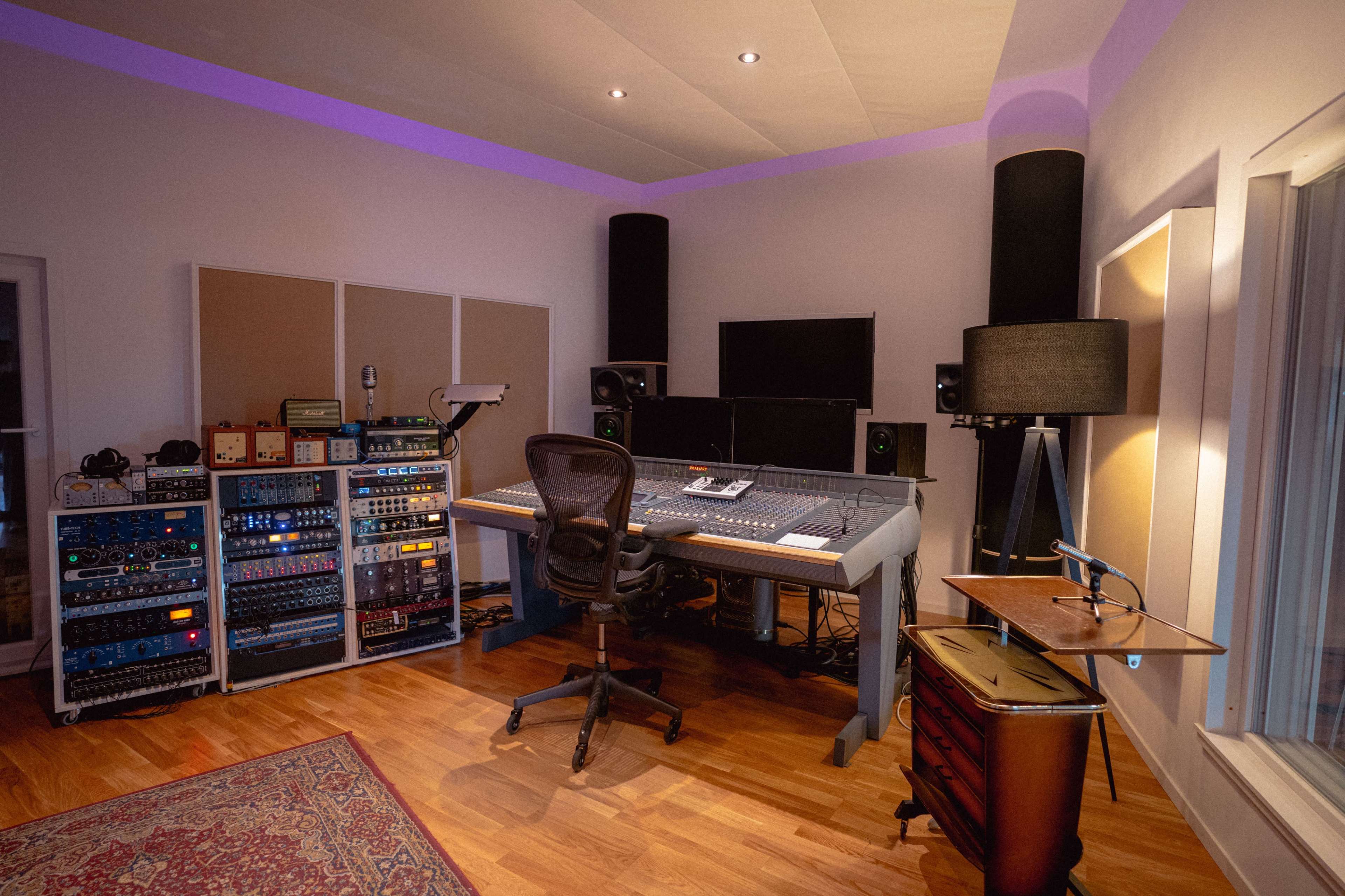 A recording studio featuring a mixing console, audio equipment racks, and a desk with studio monitors and a comfortable chair.