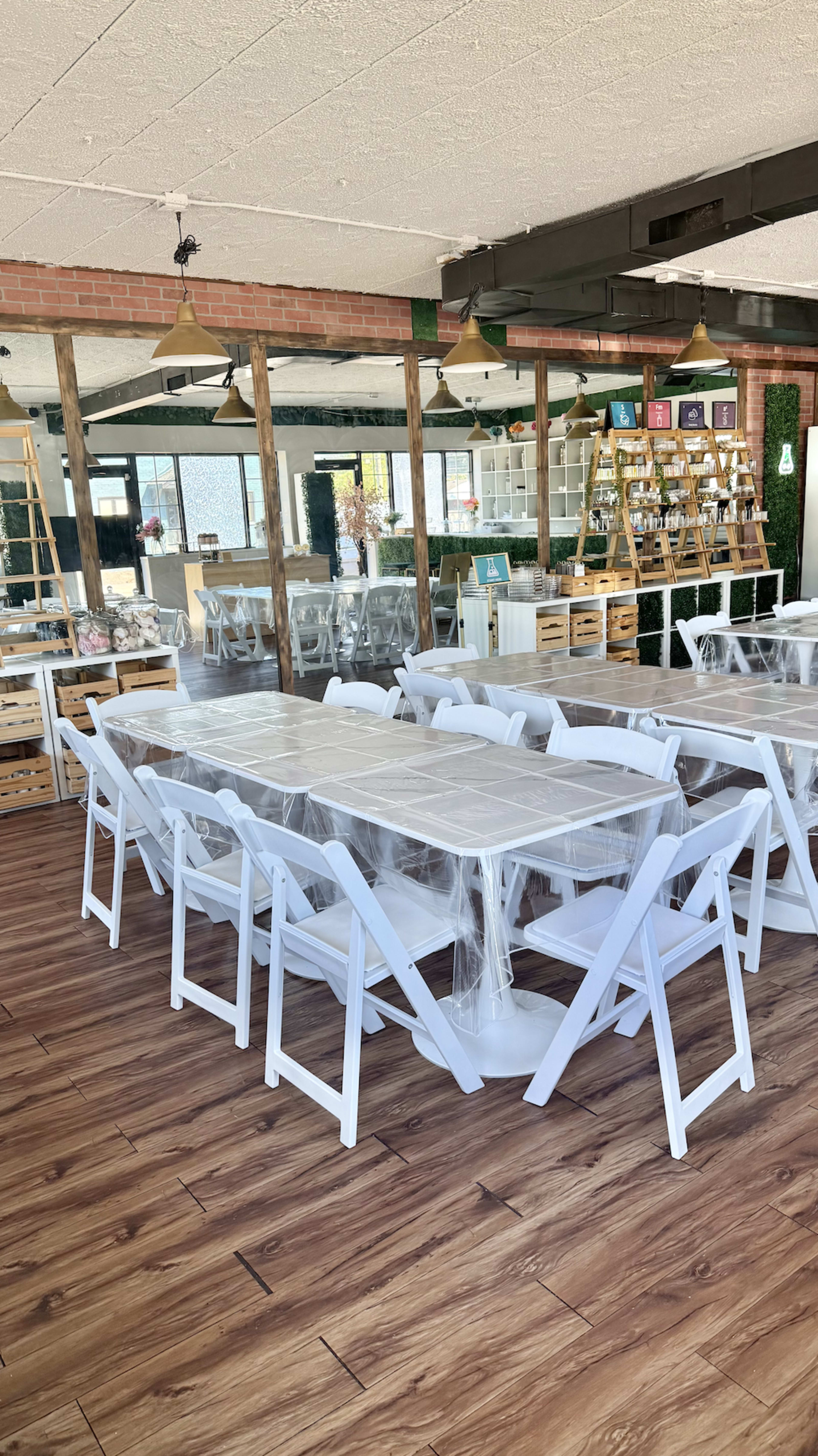 A spacious dining area features multiple white folding tables and chairs, arranged neatly on a wooden floor with natural light coming through large windows.
