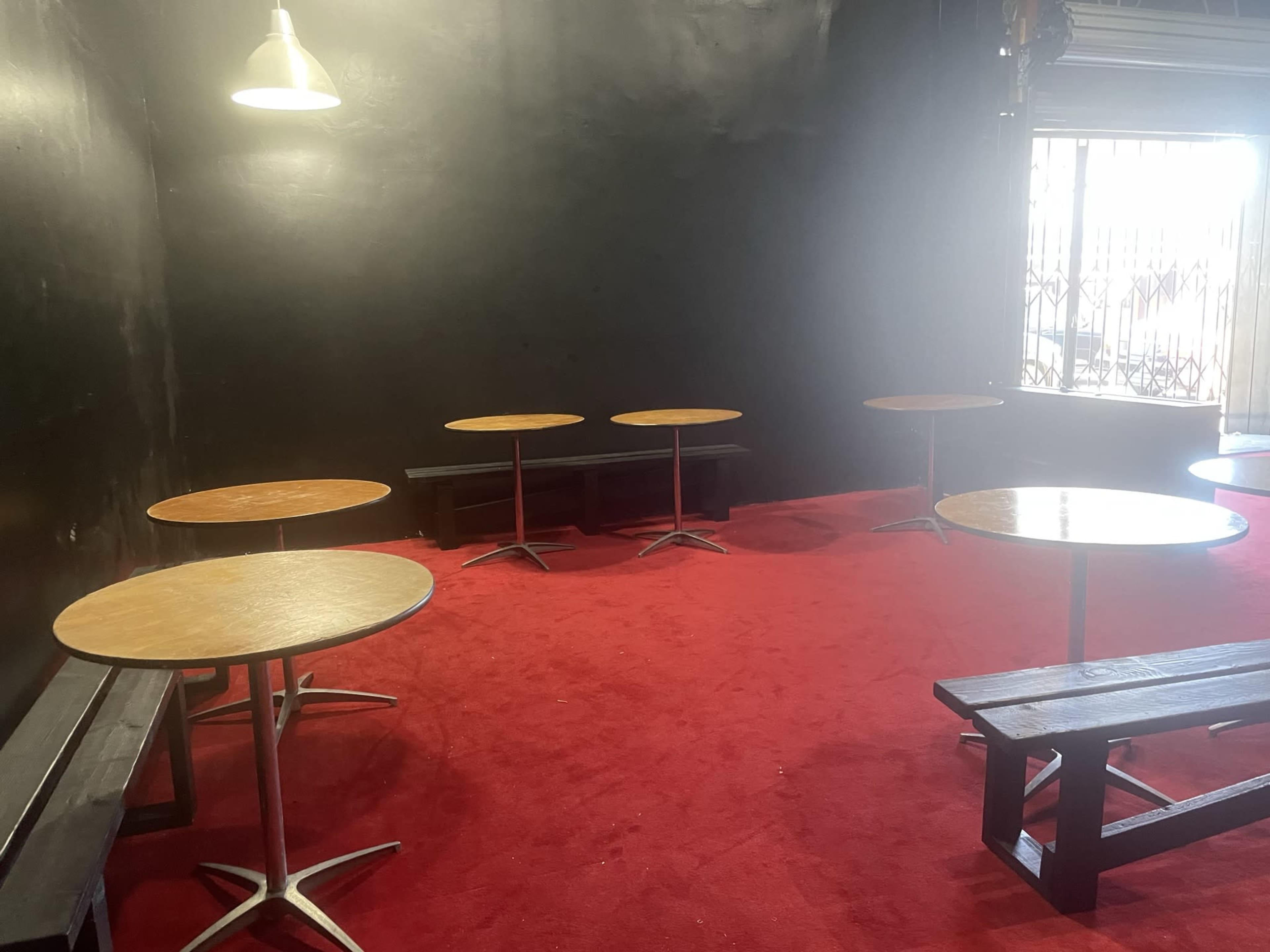 A room with several round wooden tables arranged on a red carpeted floor, accompanied by black benches near the walls and dim lighting from a hanging lamp.
