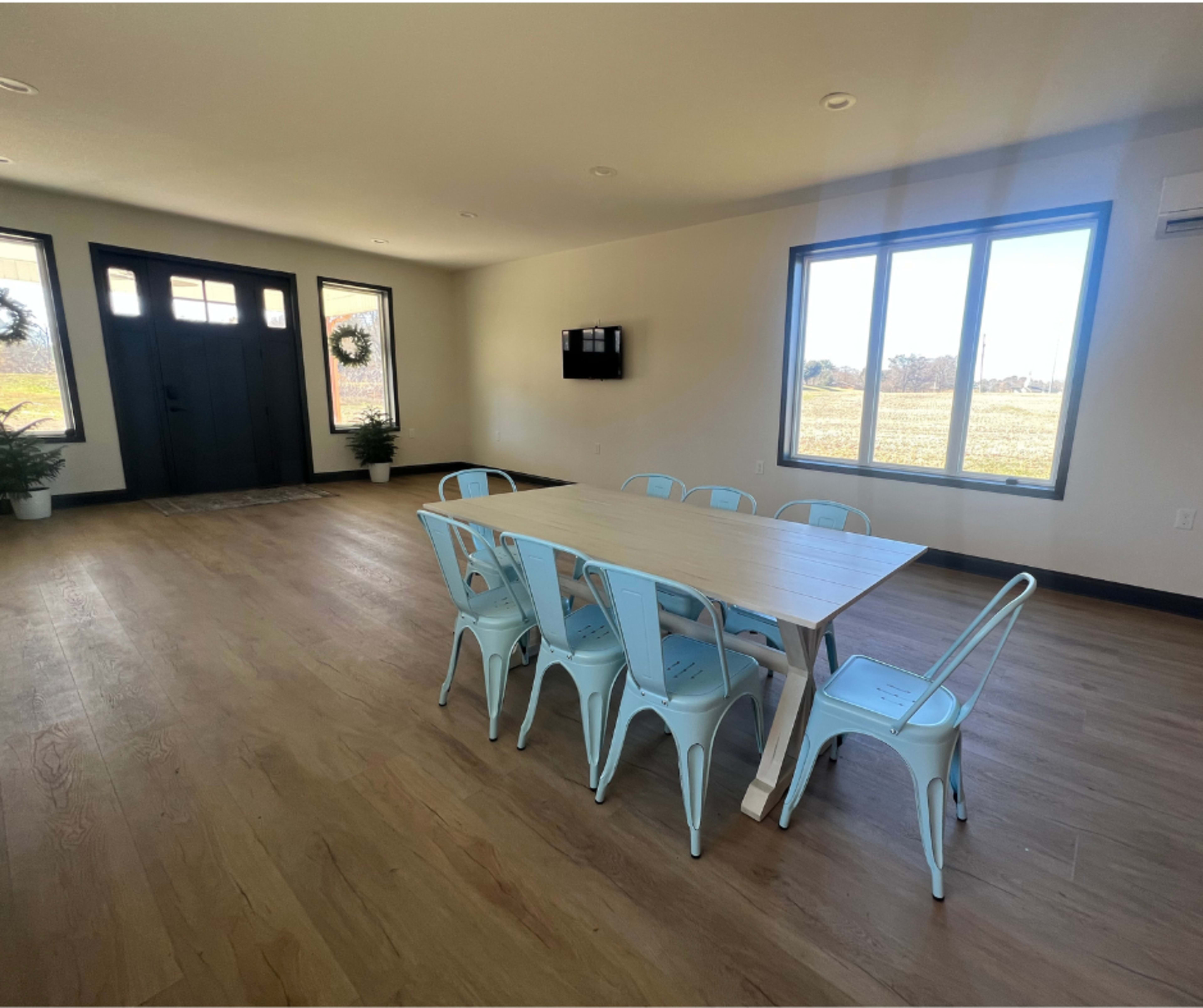 A spacious room features a wooden dining table surrounded by metal chairs, with large windows and a front door visible in the background.
