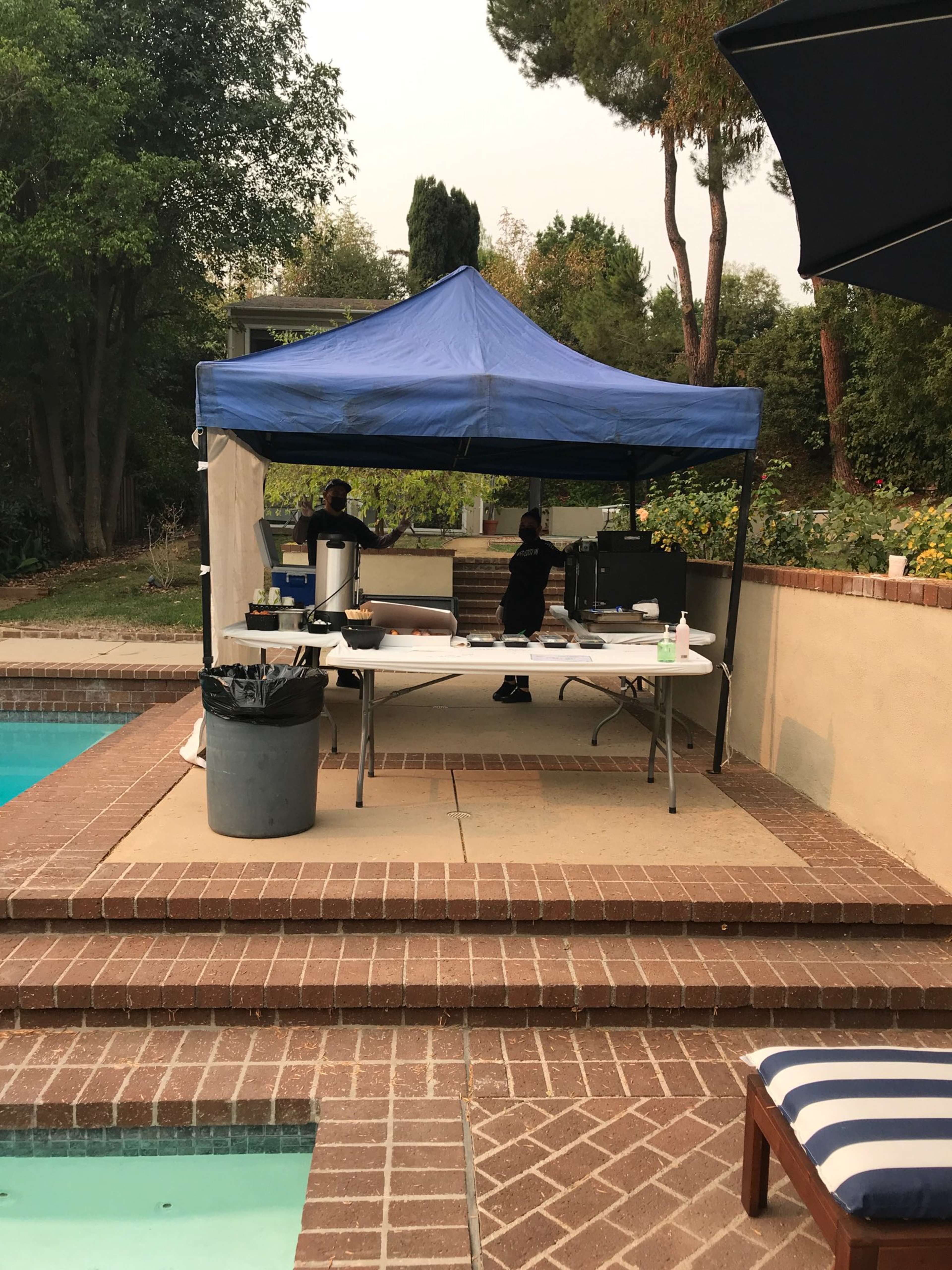 A blue canopy covers a table with cooking equipment beside a swimming pool in a landscaped backyard.