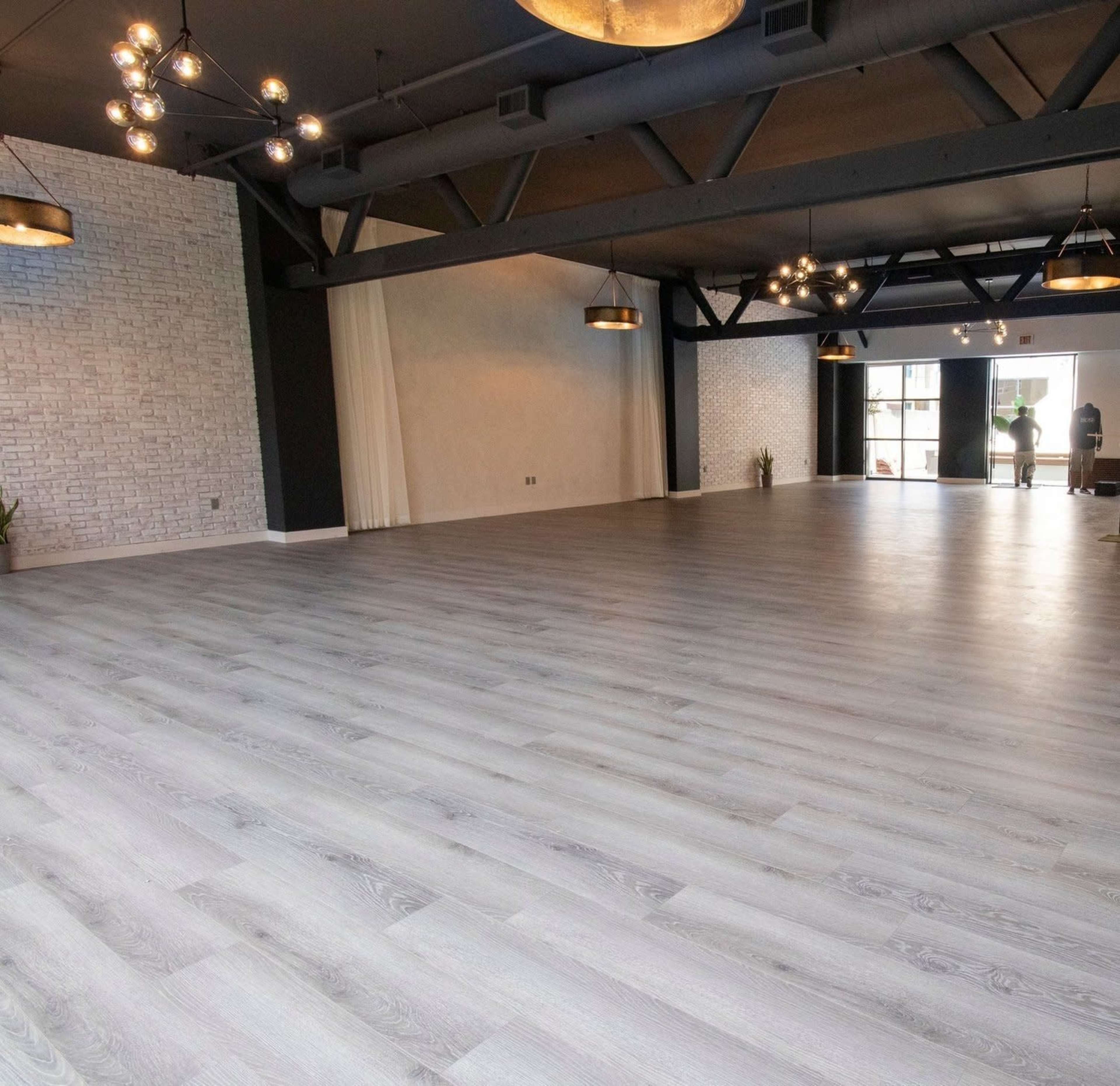 A spacious interior with grey wooden flooring, exposed beams, and large windows allowing natural light to enter.