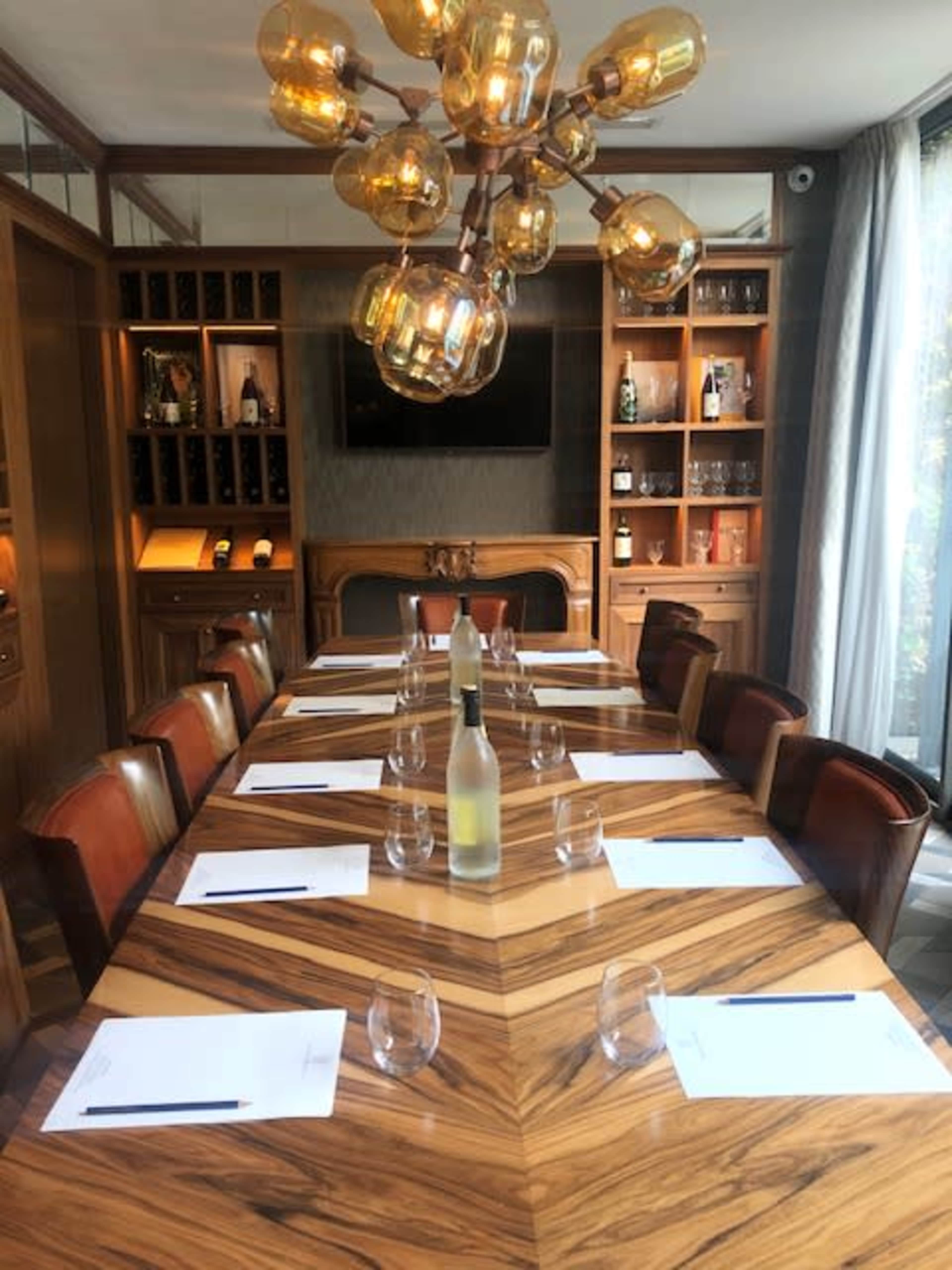 A long wooden conference table is set with glasses and notepads, surrounded by leather chairs, with a modern chandelier overhead and shelves displaying bottles in the background.