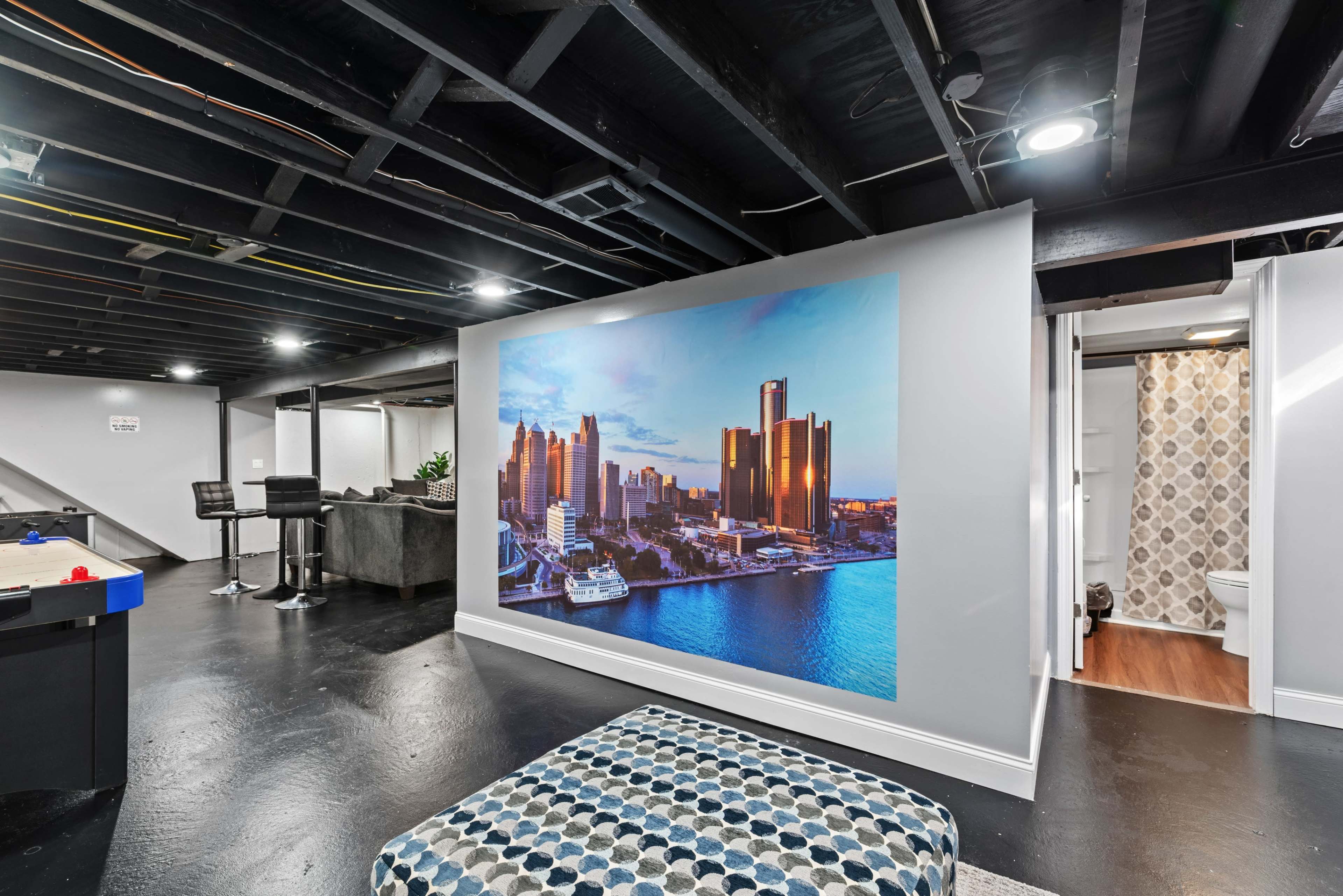 A basement room featuring a large mural of a city skyline on one wall, with modern furnishings and a small bathroom area visible in the background.