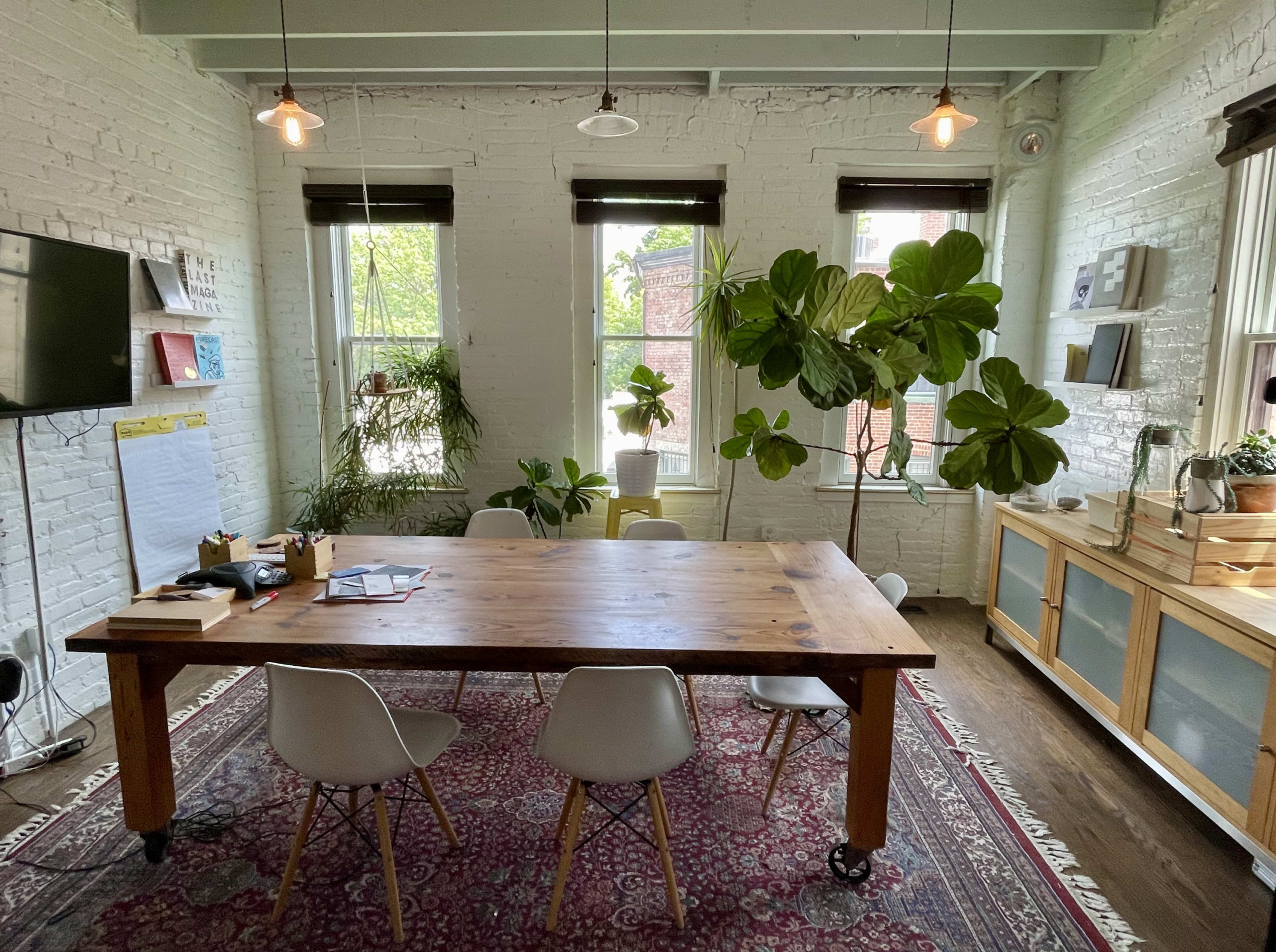 The image shows a bright, modern meeting room with a wooden table surrounded by chairs, large windows, and potted plants.