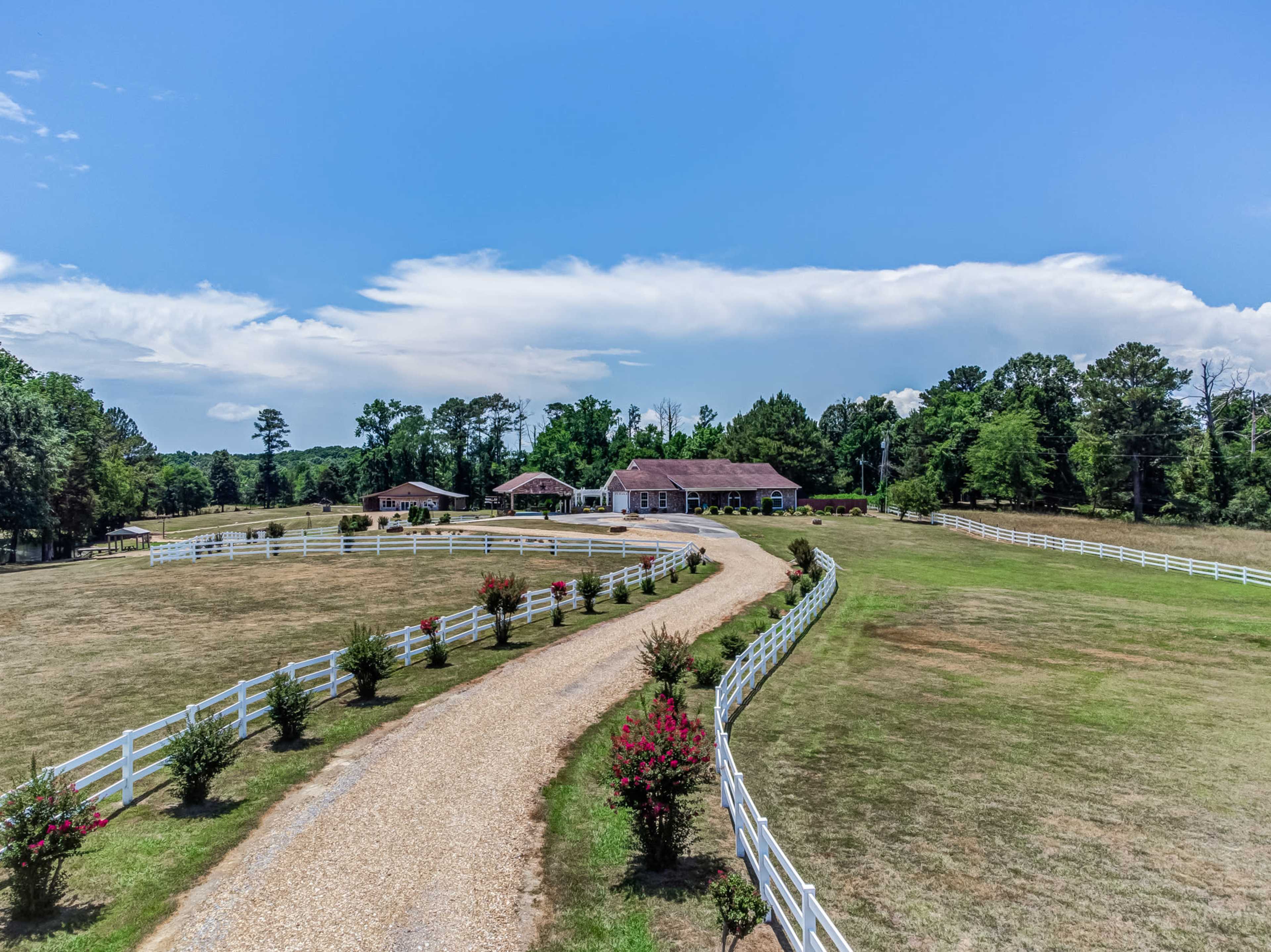 A winding gravel driveway leads to a house surrounded by green lawns and white fences under a blue sky.