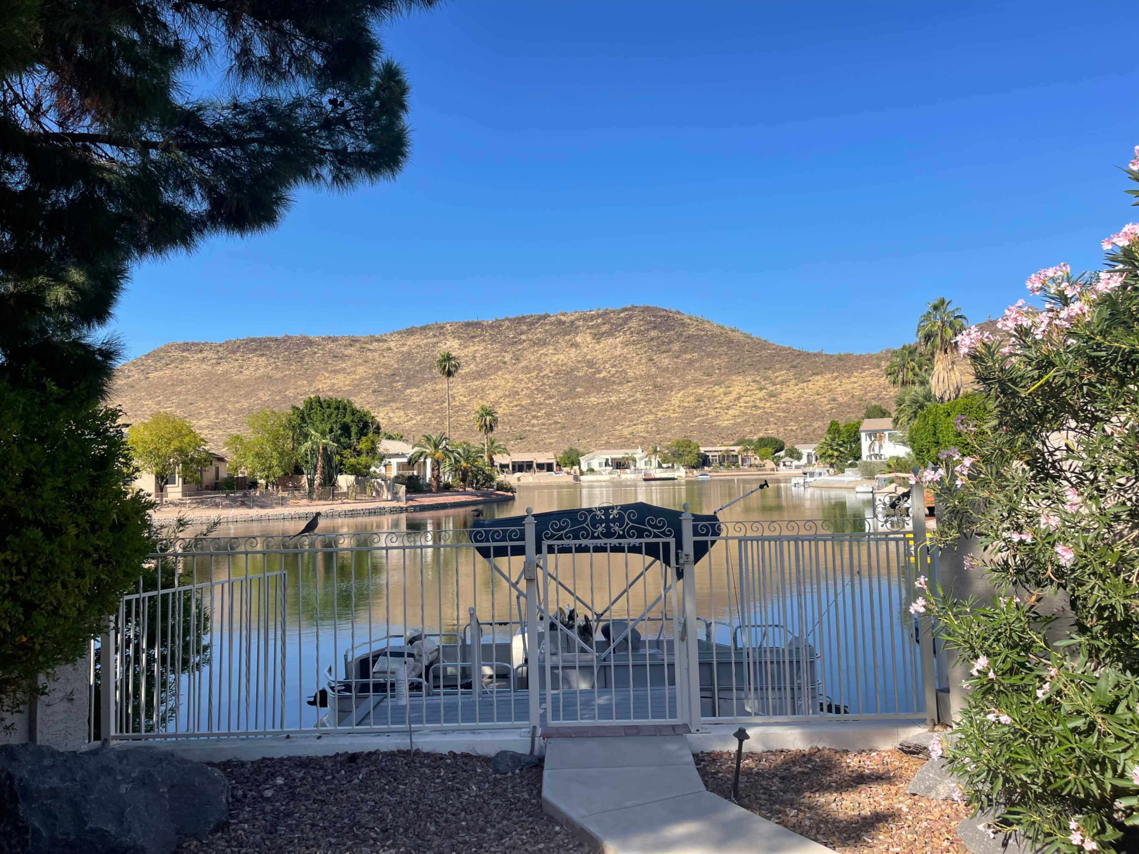The image shows a serene lakeside view framed by a white gated entrance, with a houseboat moored along the water's edge and a mountain in the background.