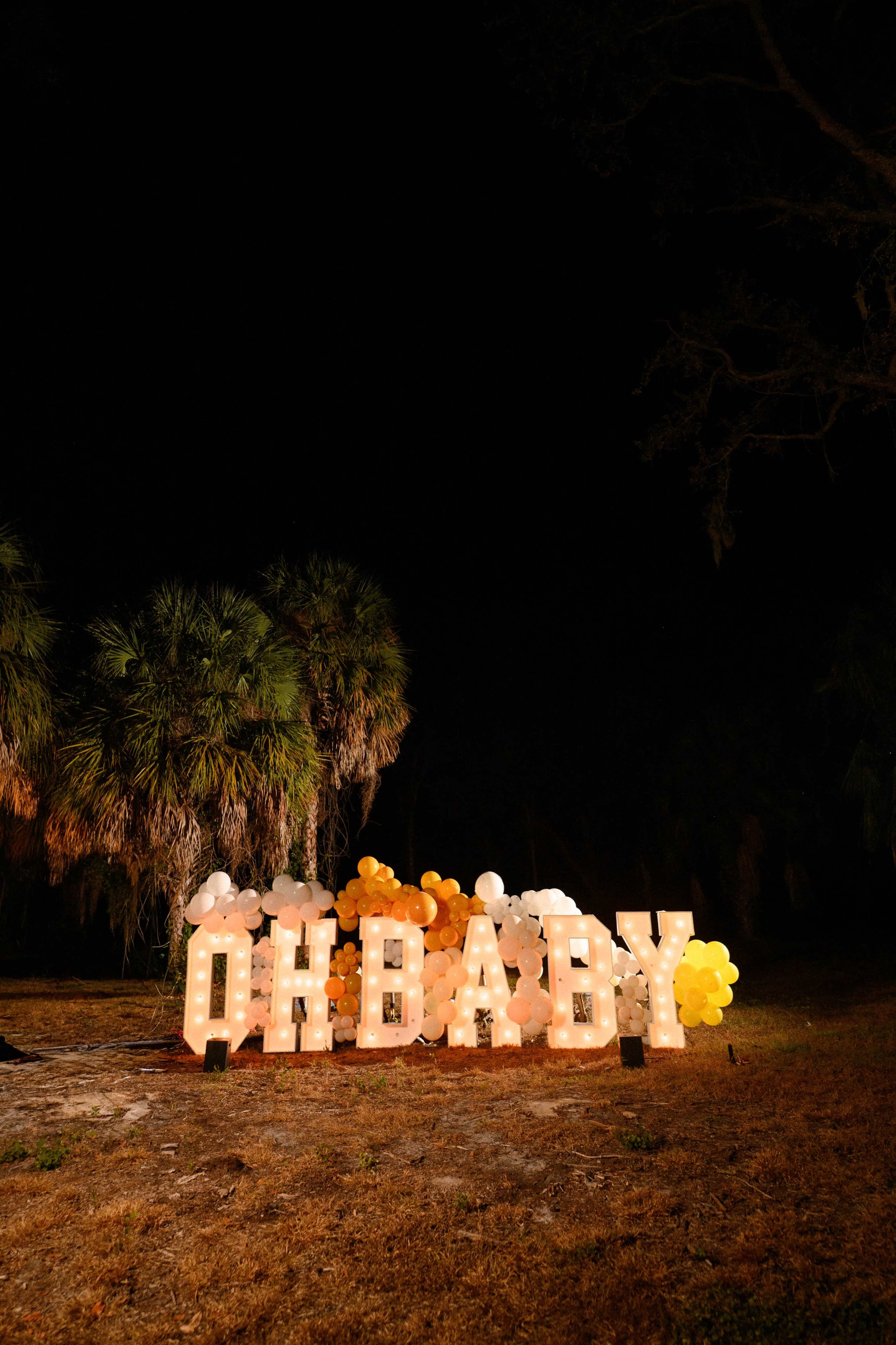 The scene features oversized illuminated letters spelling "OH BABY," surrounded by colorful balloons, set against a dark backdrop with palm trees.