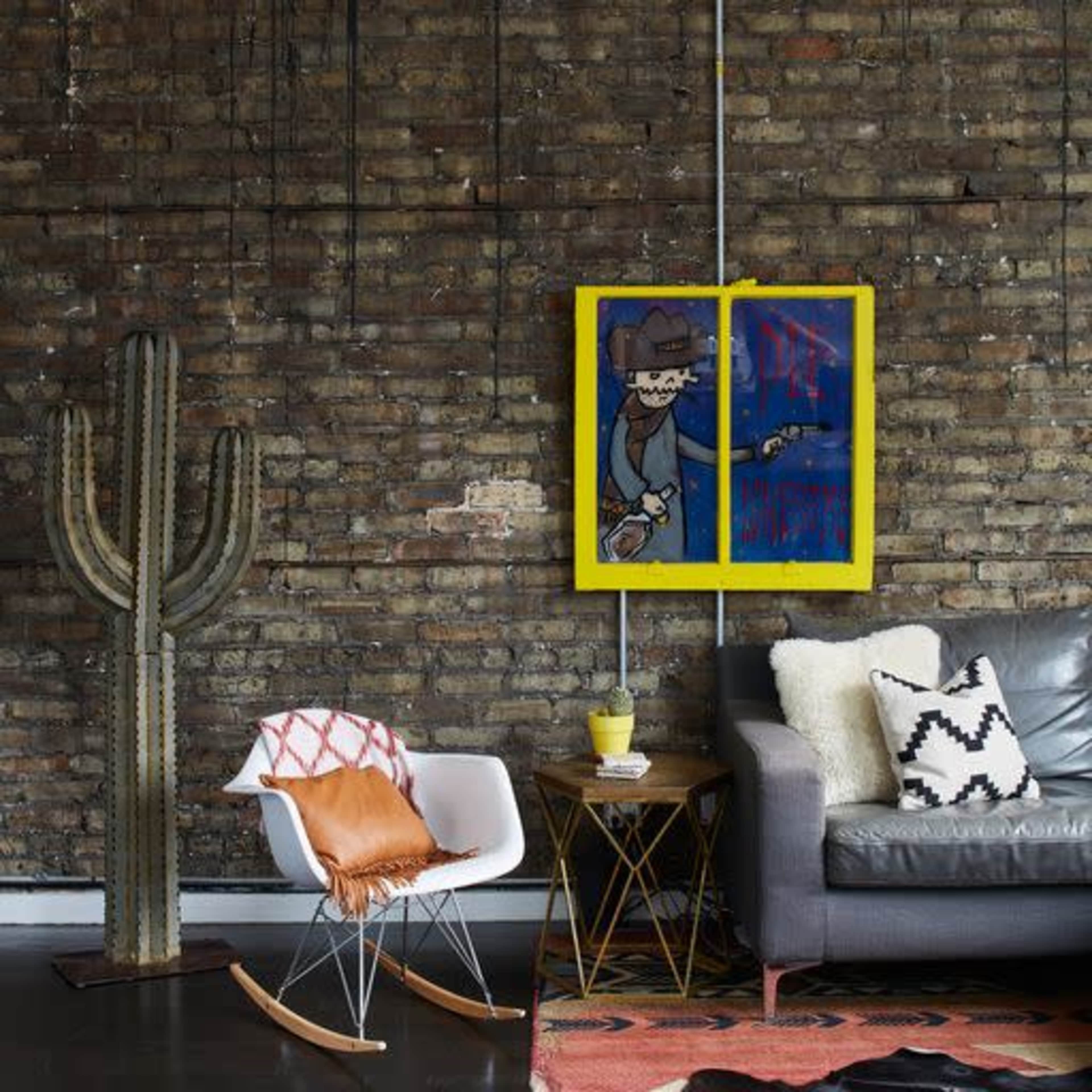The image features a modern living space with a cactus, a stylish chair, a table, and a sofa against a brick wall adorned with colorful artwork.