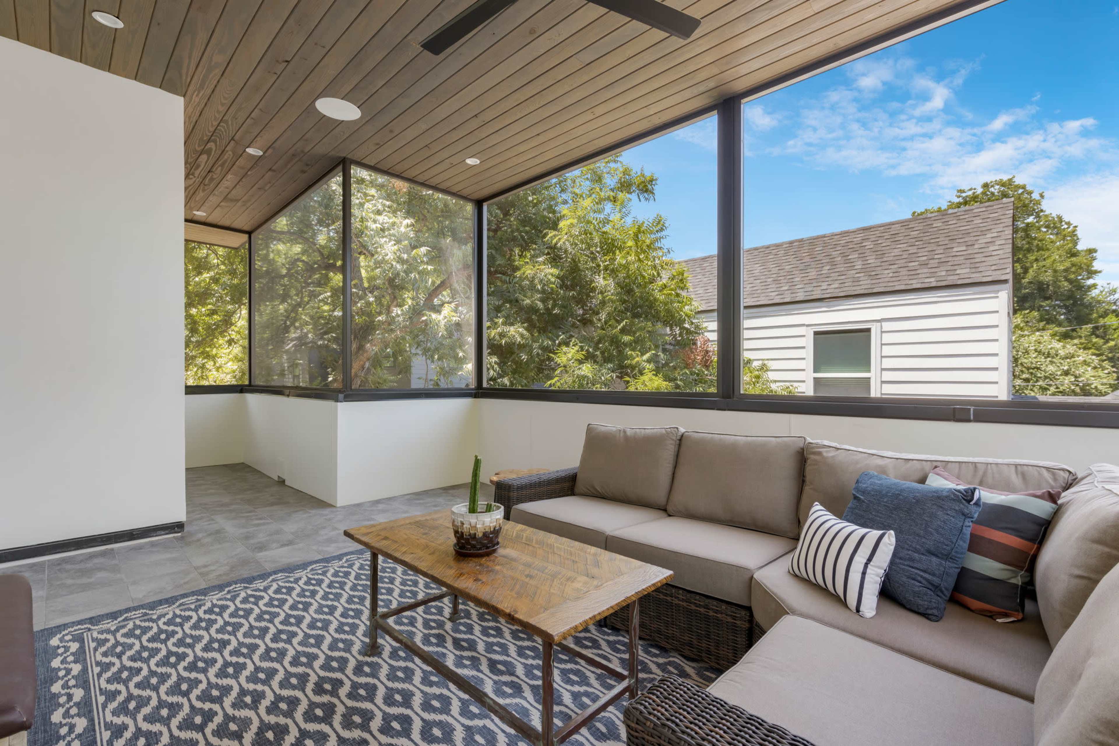 The image shows a screened-in porch featuring a comfortable sectional sofa, a wooden coffee table, and large windows that provide views of greenery outside.