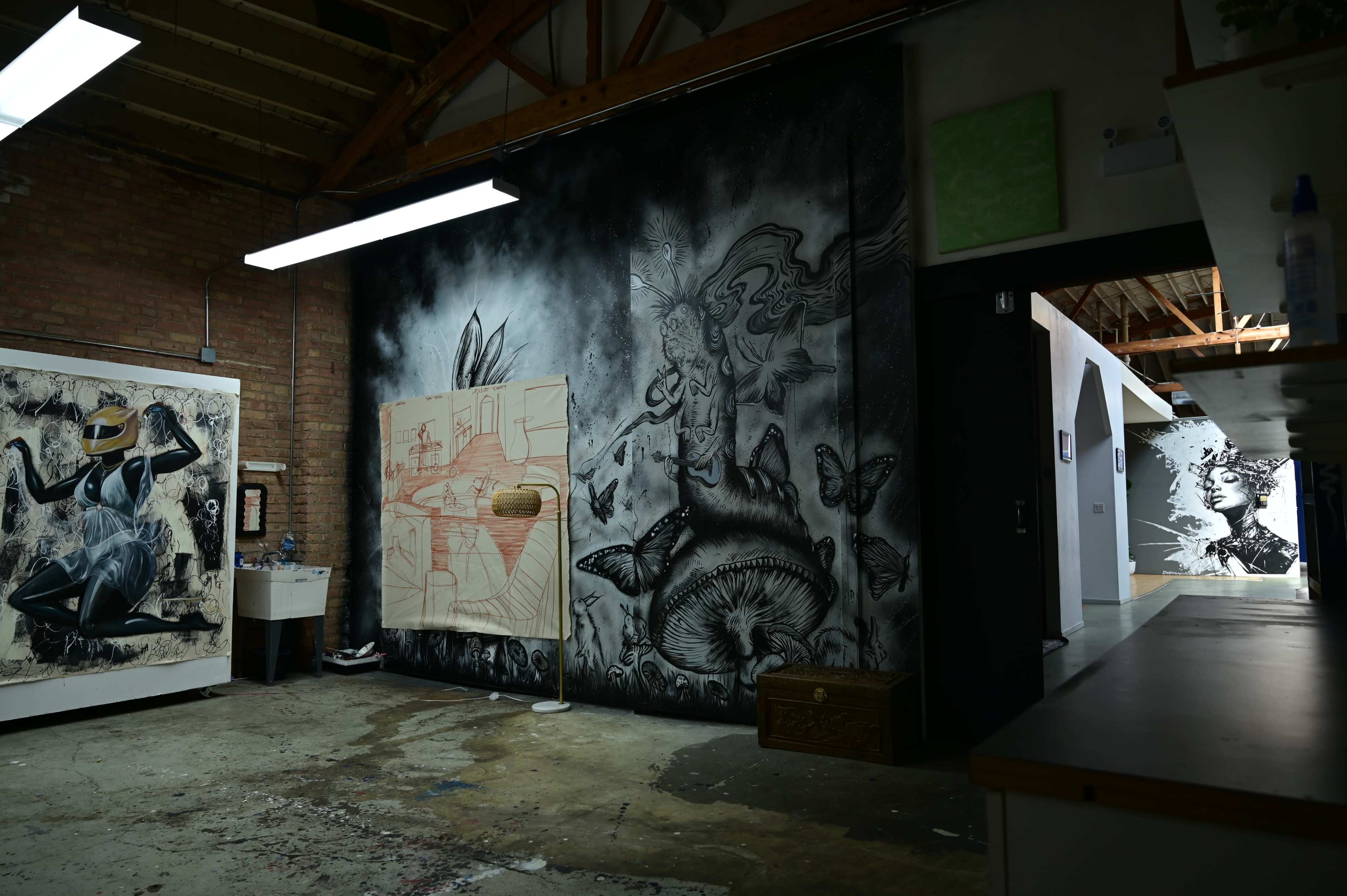 The image shows an art studio with a large mural on one wall, featuring a figure amidst butterflies and mushrooms, and various artworks displayed around the space.