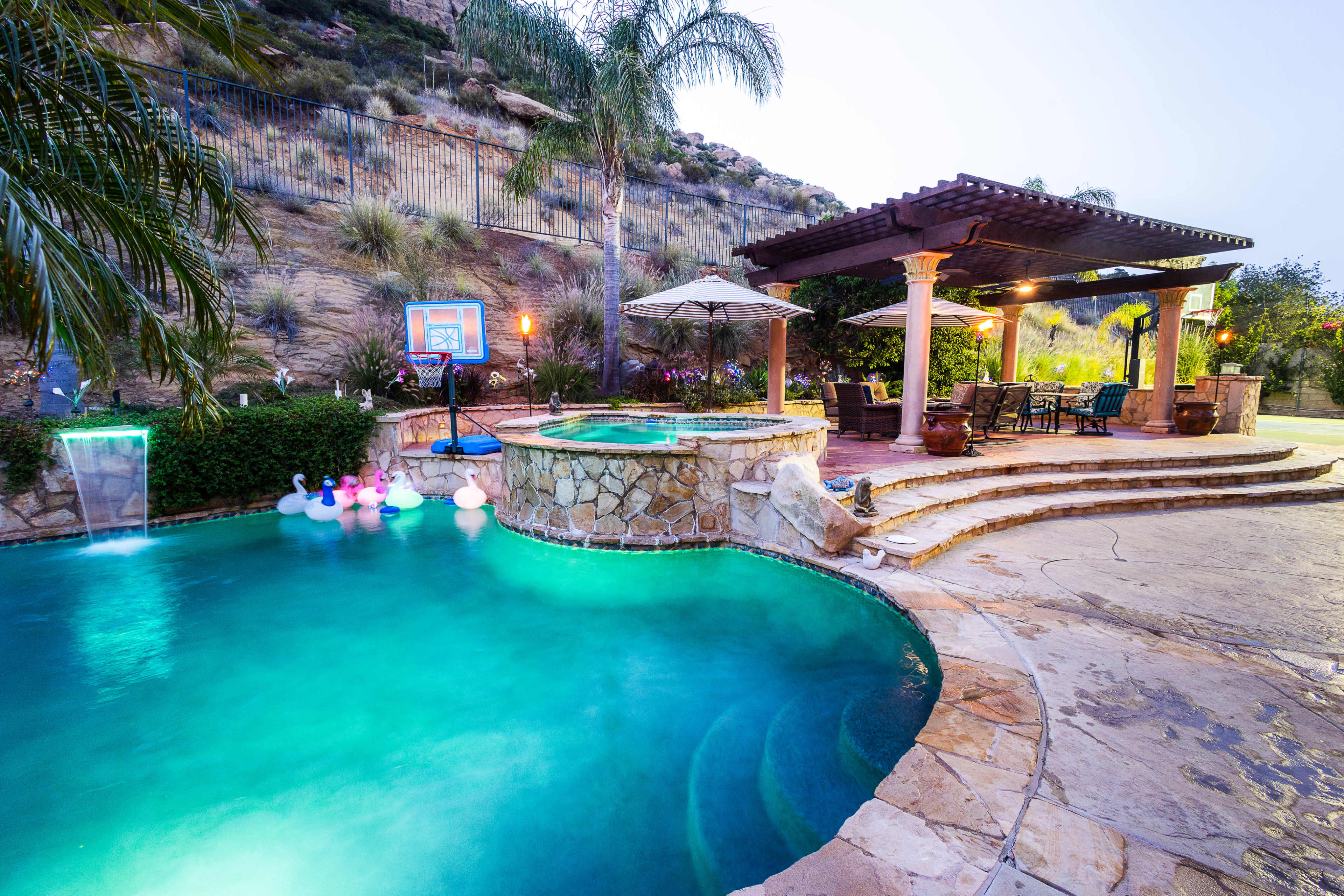 The image shows a landscaped backyard with a swimming pool, a spa area, a shaded patio, and decorative pool floats.