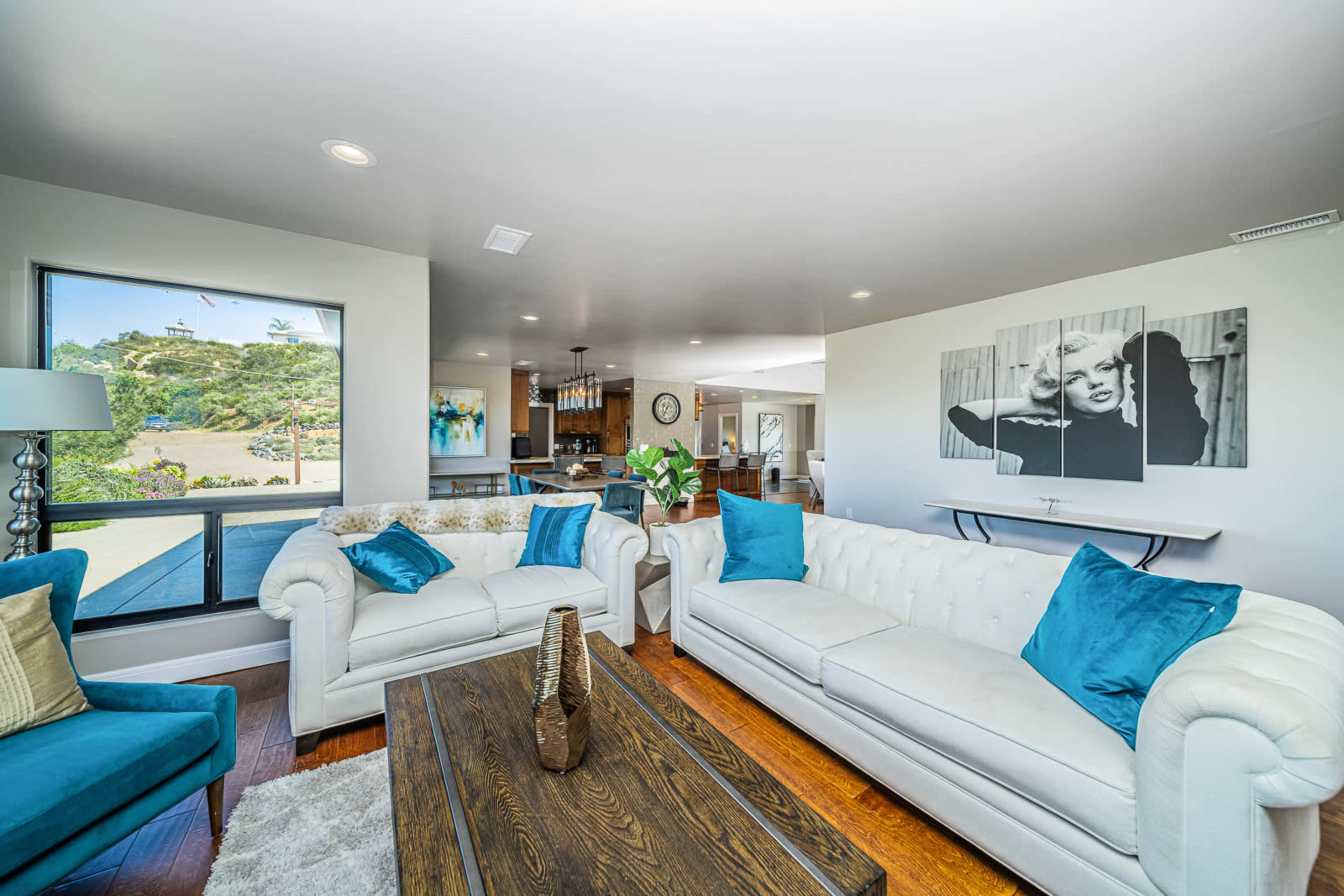 A modern living room features two white sofas with blue cushions, a wooden coffee table, and a large window overlooking a garden.
