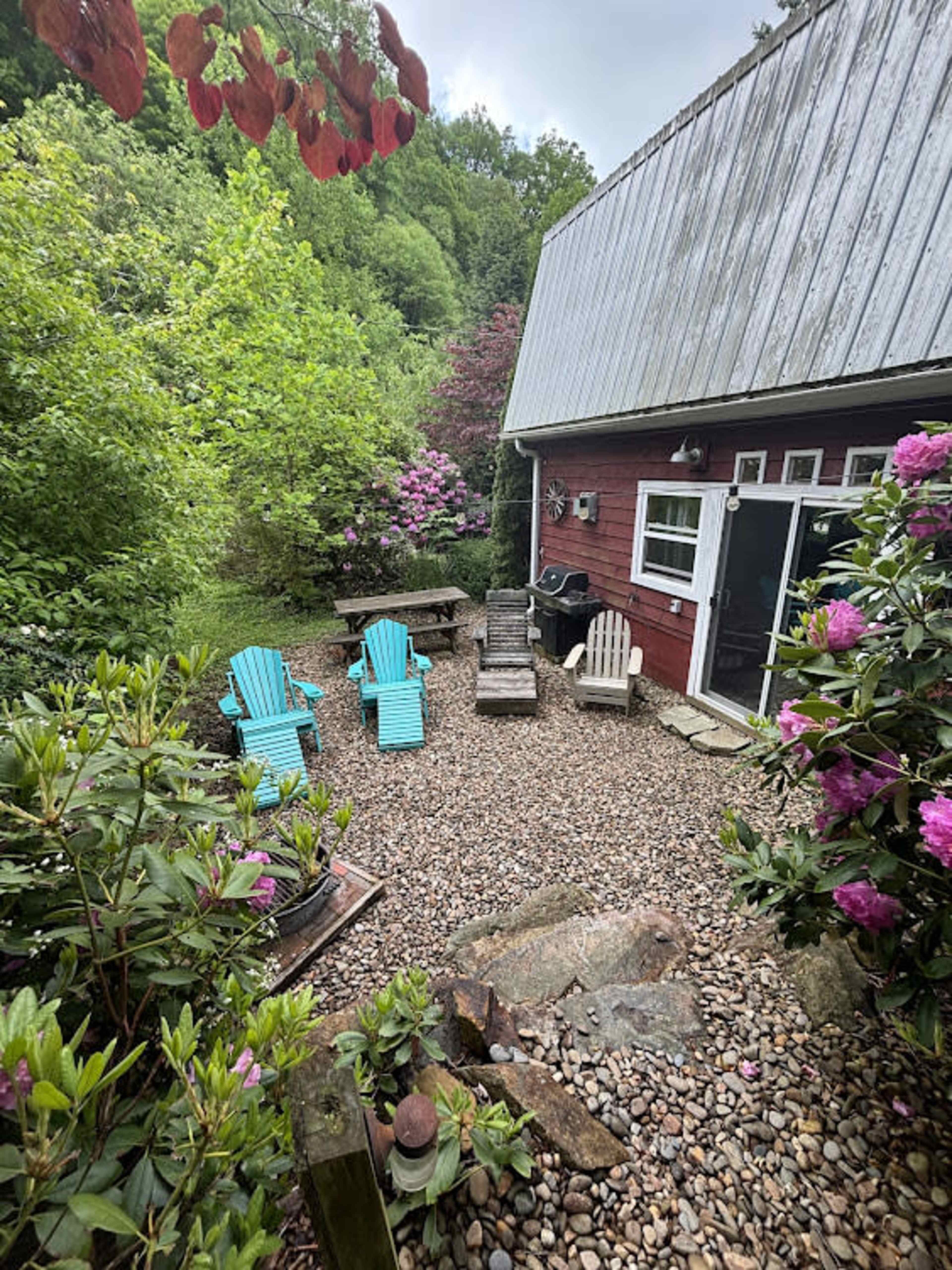 A cozy outdoor area features colorful chairs, a gravel patio, and lush greenery surrounding a red building.