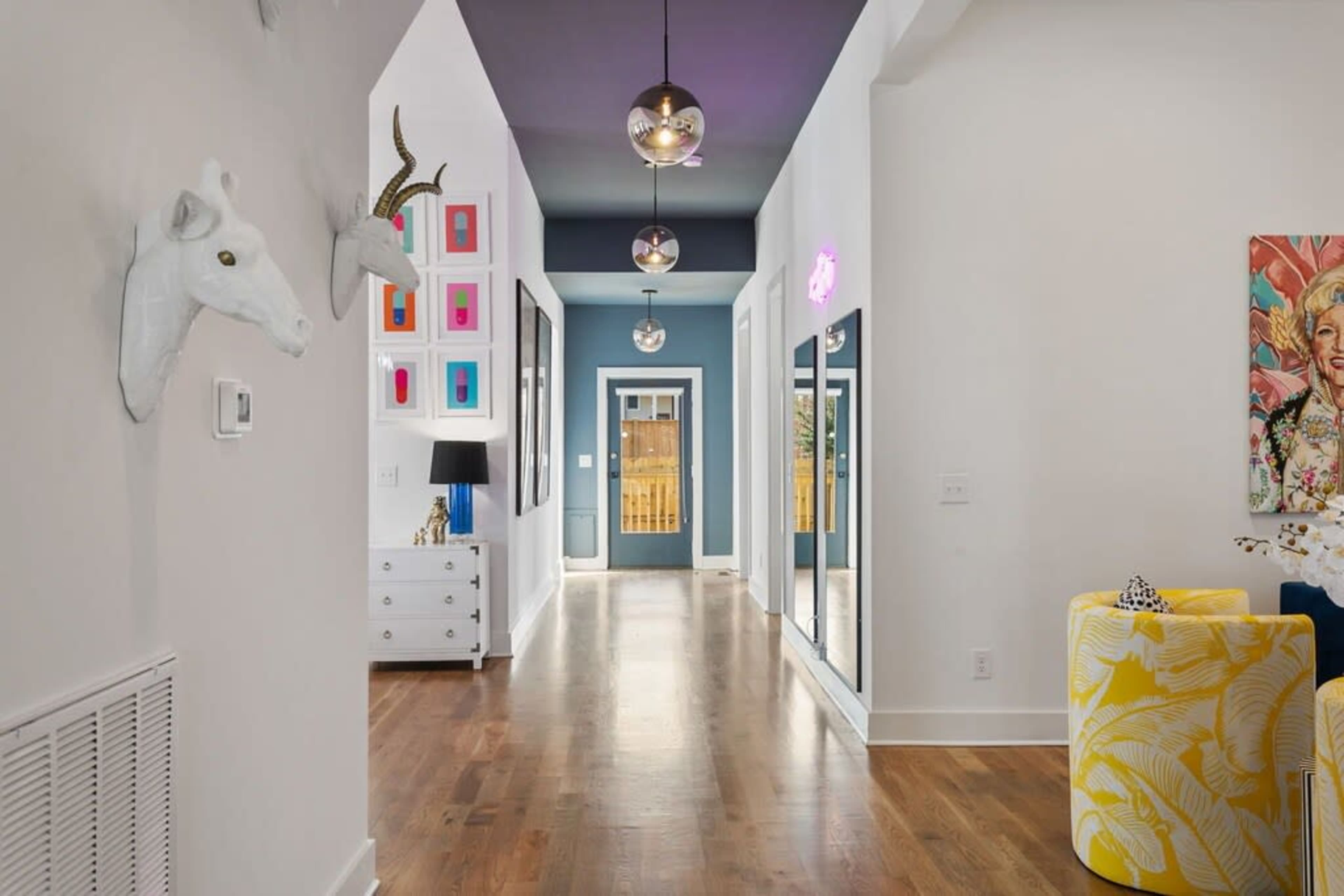 The image shows a bright hallway with dark blue walls, wooden flooring, and decorative items, including a unicorn head and a collection of colorful framed artworks.
