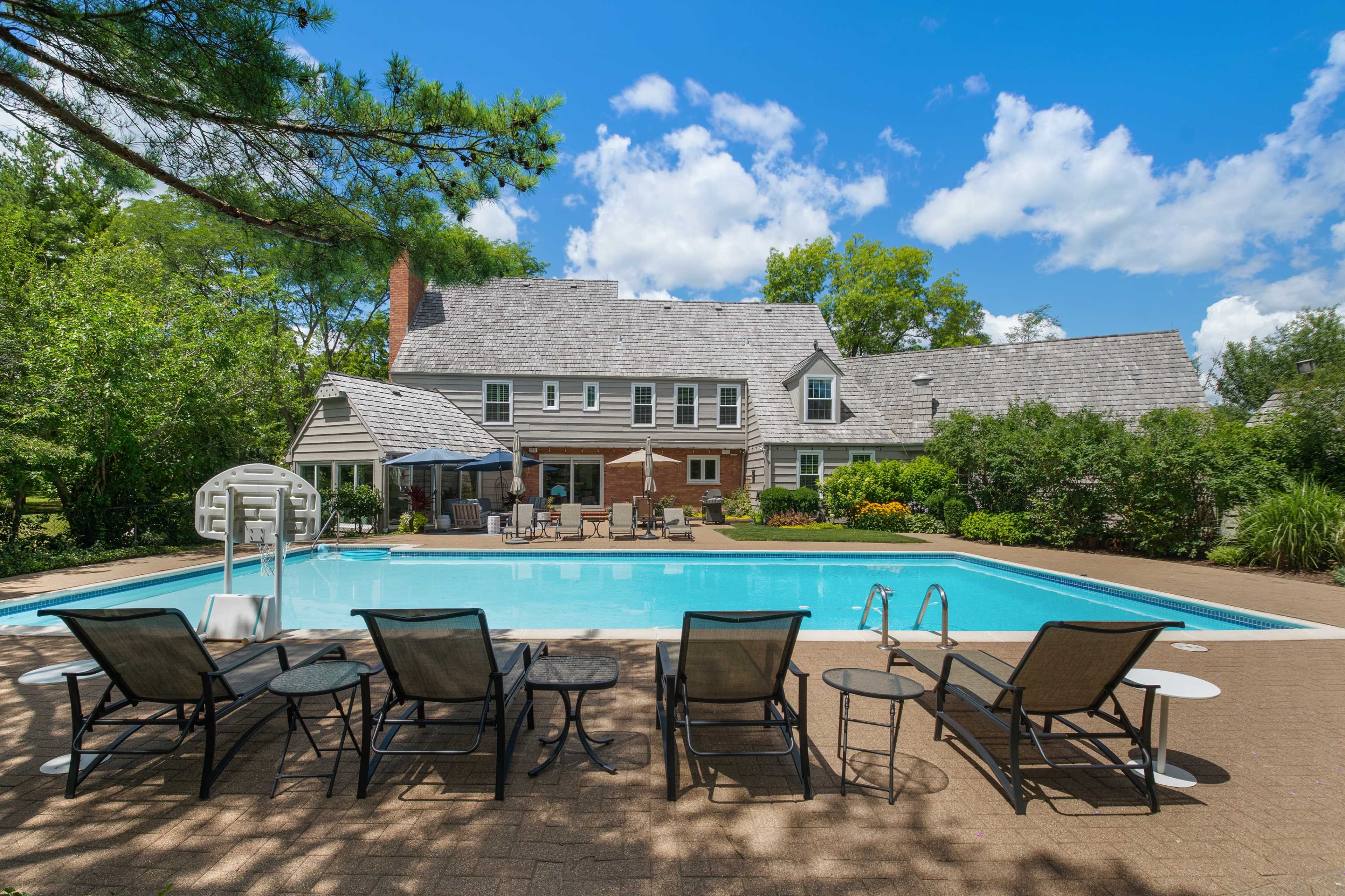A large swimming pool is surrounded by chairs and tables, with a spacious house and lush greenery in the background under a partly cloudy sky.