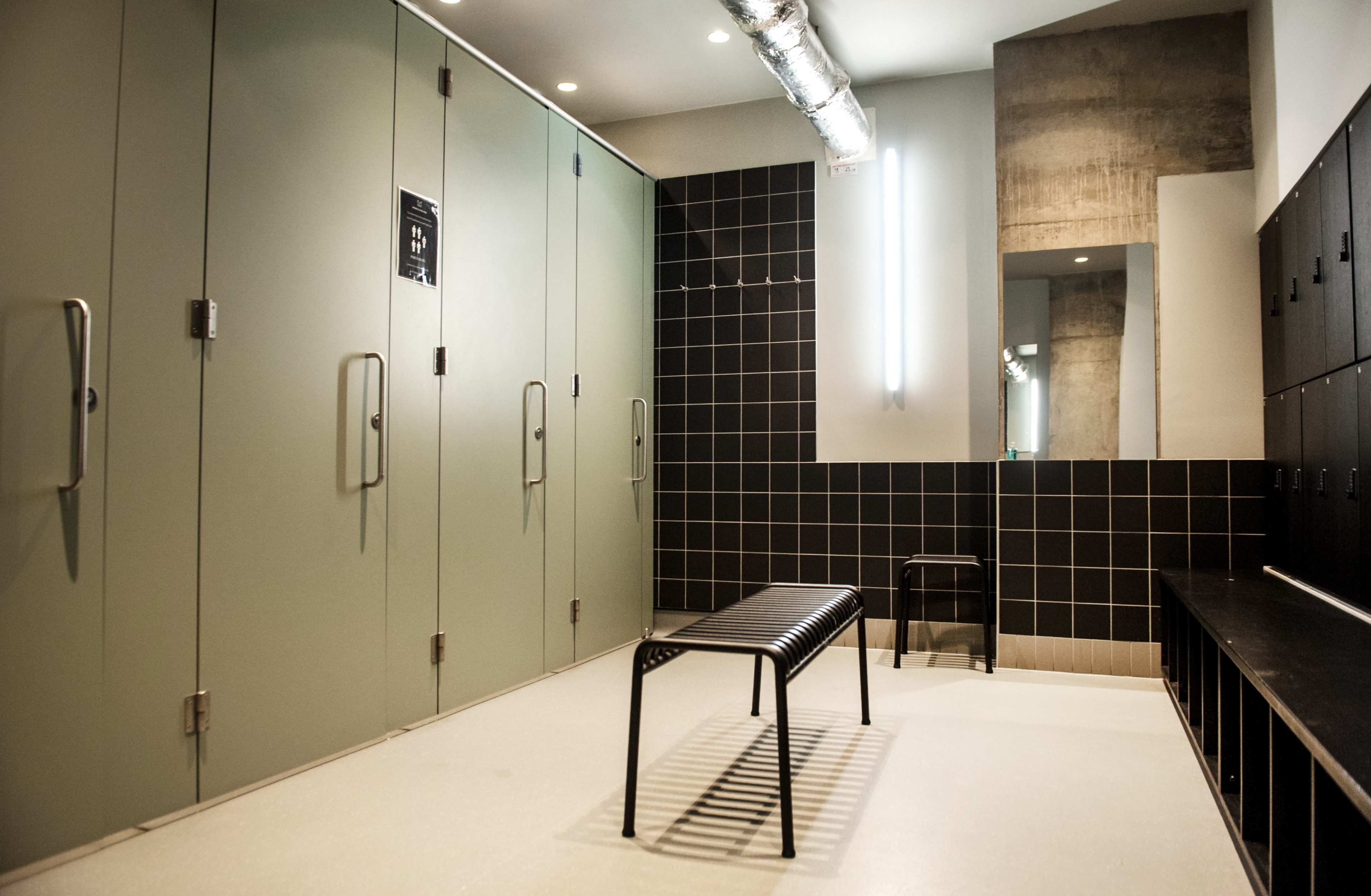 The image shows a minimalistic changing room with several closed gray lockers and a black bench against a tiled wall.