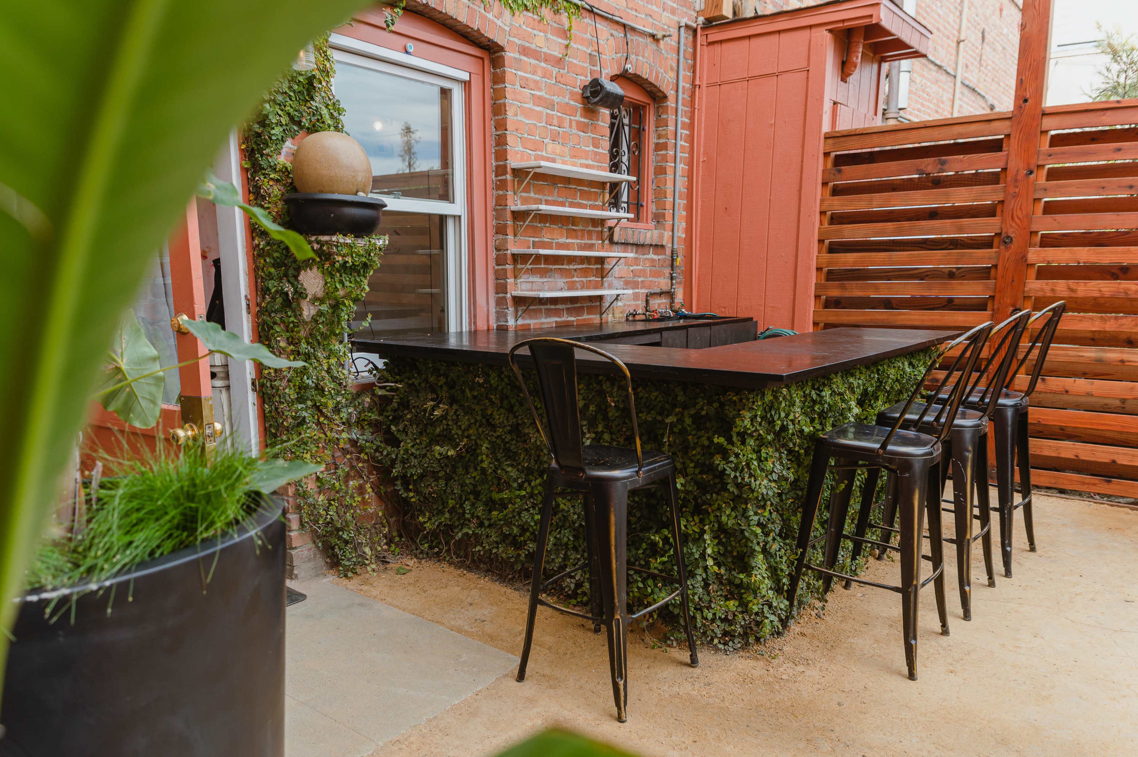 A small outdoor bar area with a black countertop and metal stools, surrounded by brick walls and greenery.