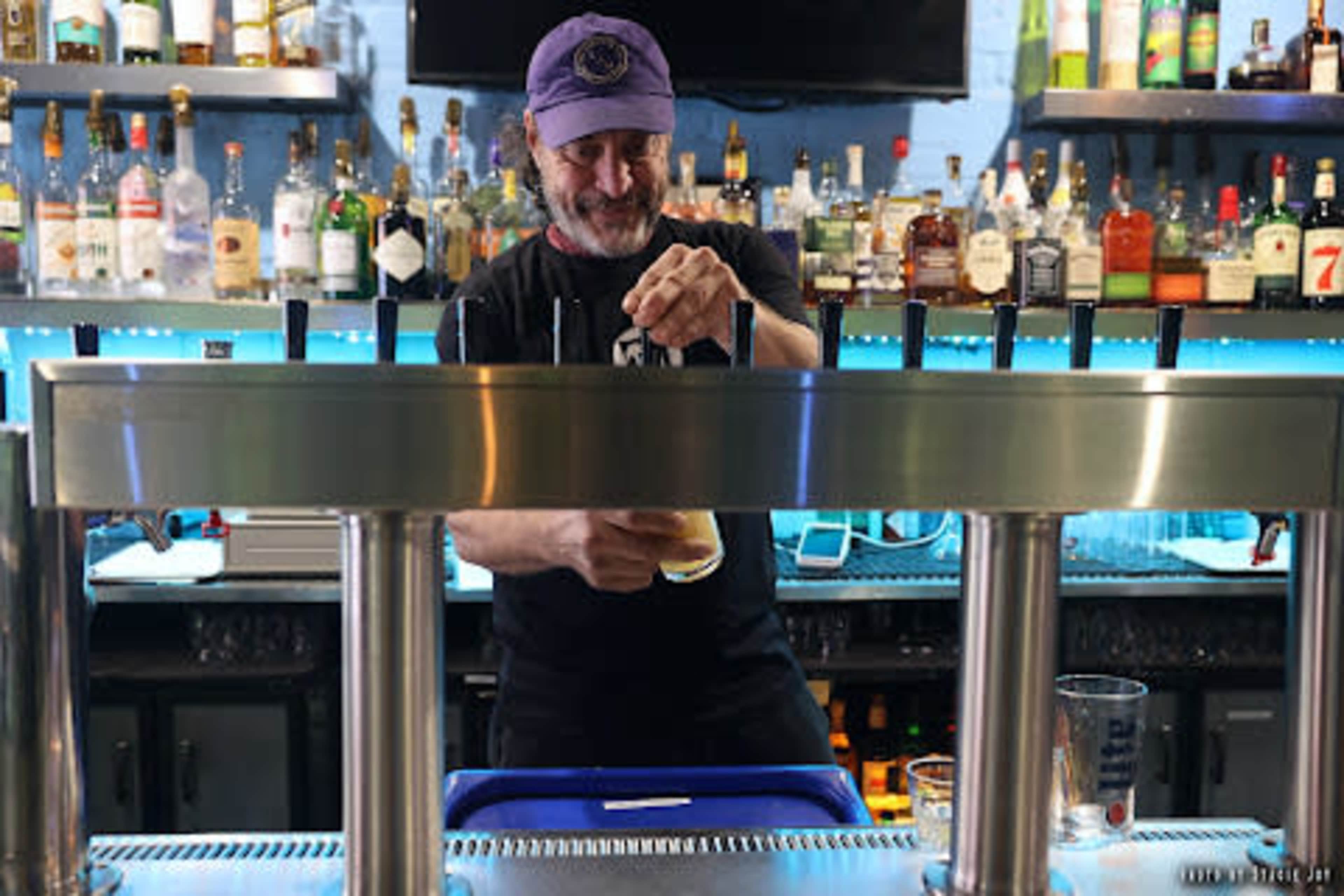 A bartender pours a drink from a tap at a bar lined with various bottles of liquor.