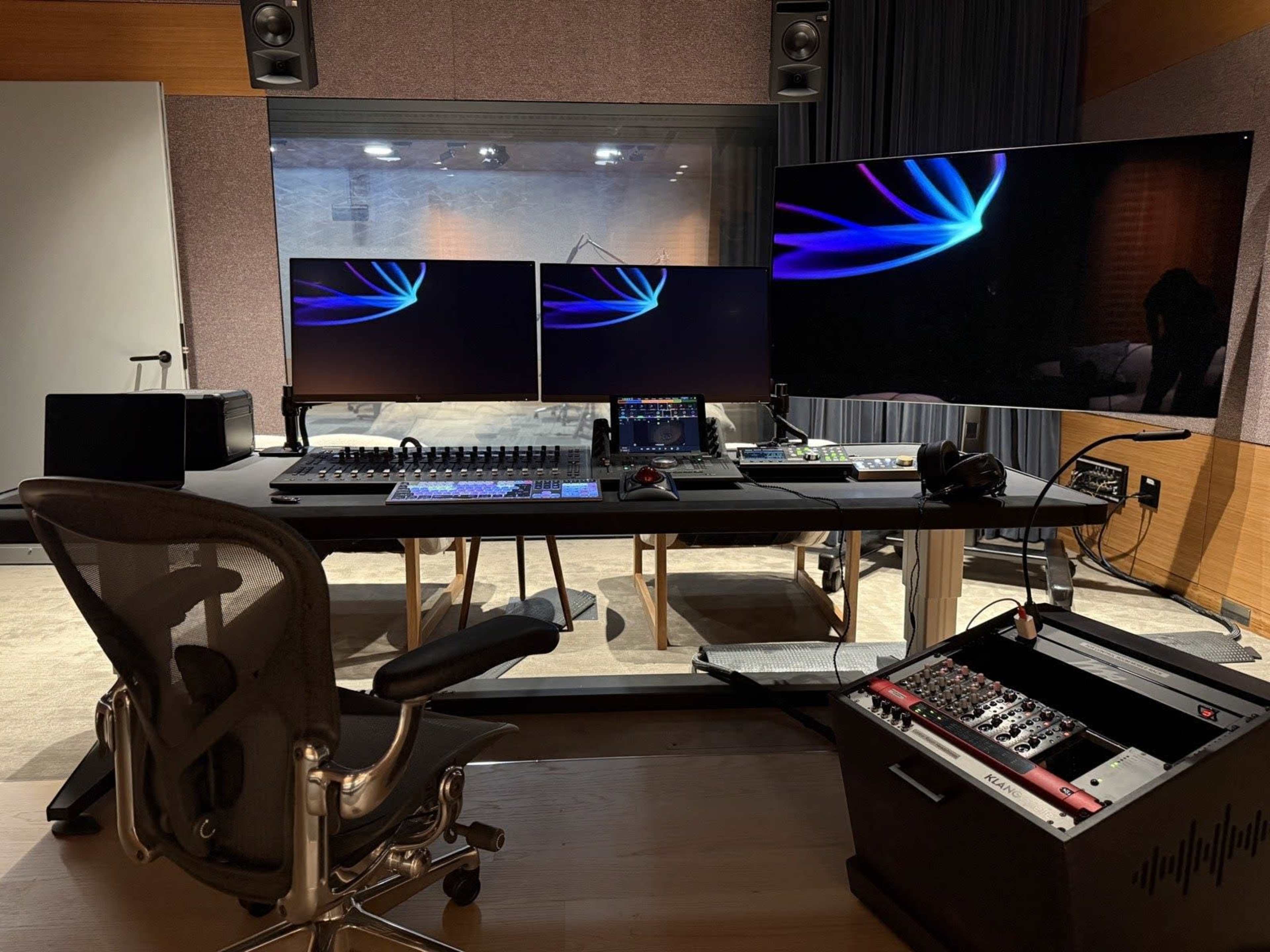 The image shows a modern audio or video editing studio featuring a large desk with dual computer monitors, a mixing console, and various electronic equipment.