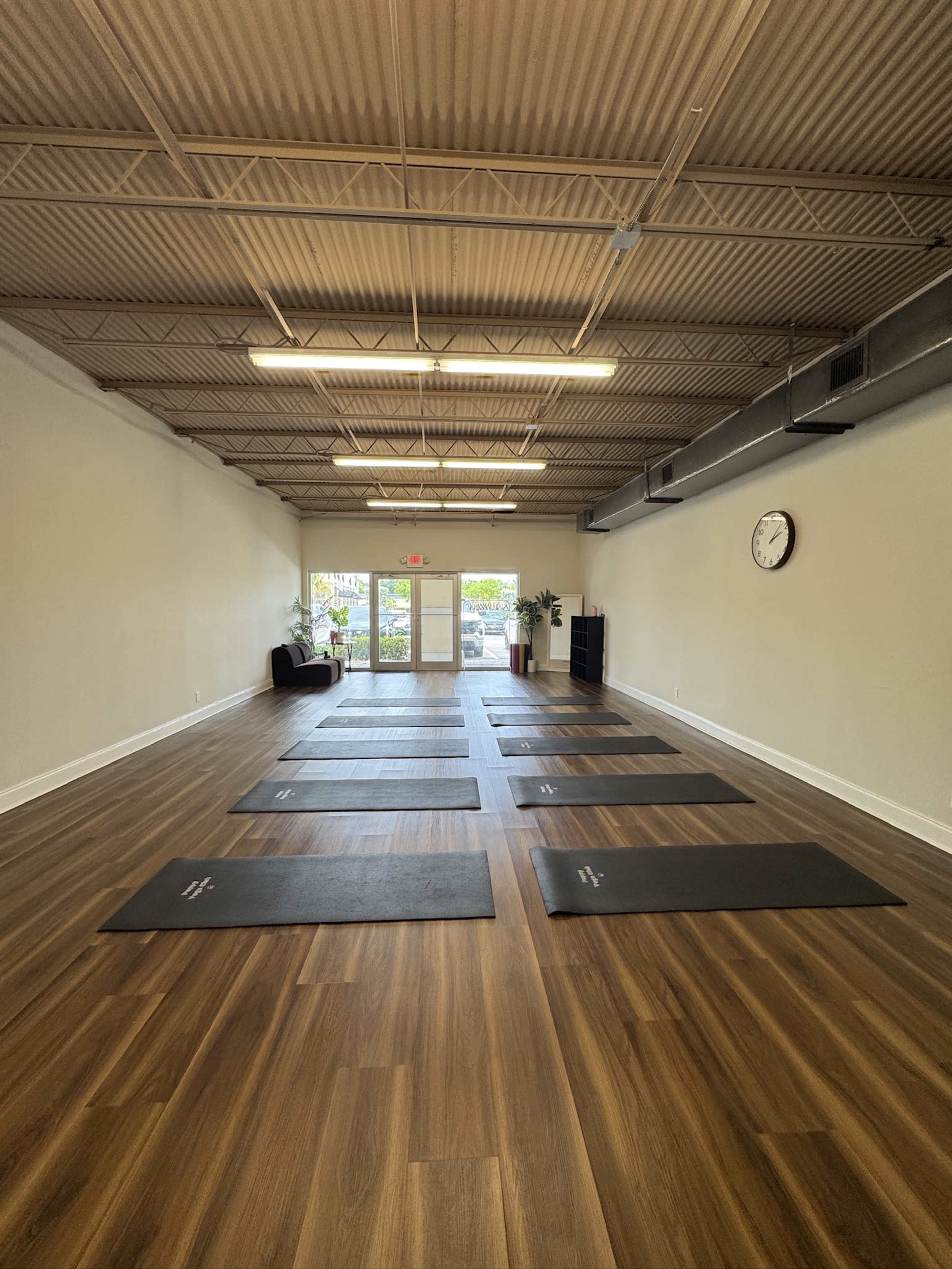 The image shows a spacious yoga studio with wooden floors, eight yoga mats arranged neatly, and a wall clock visible in the background.