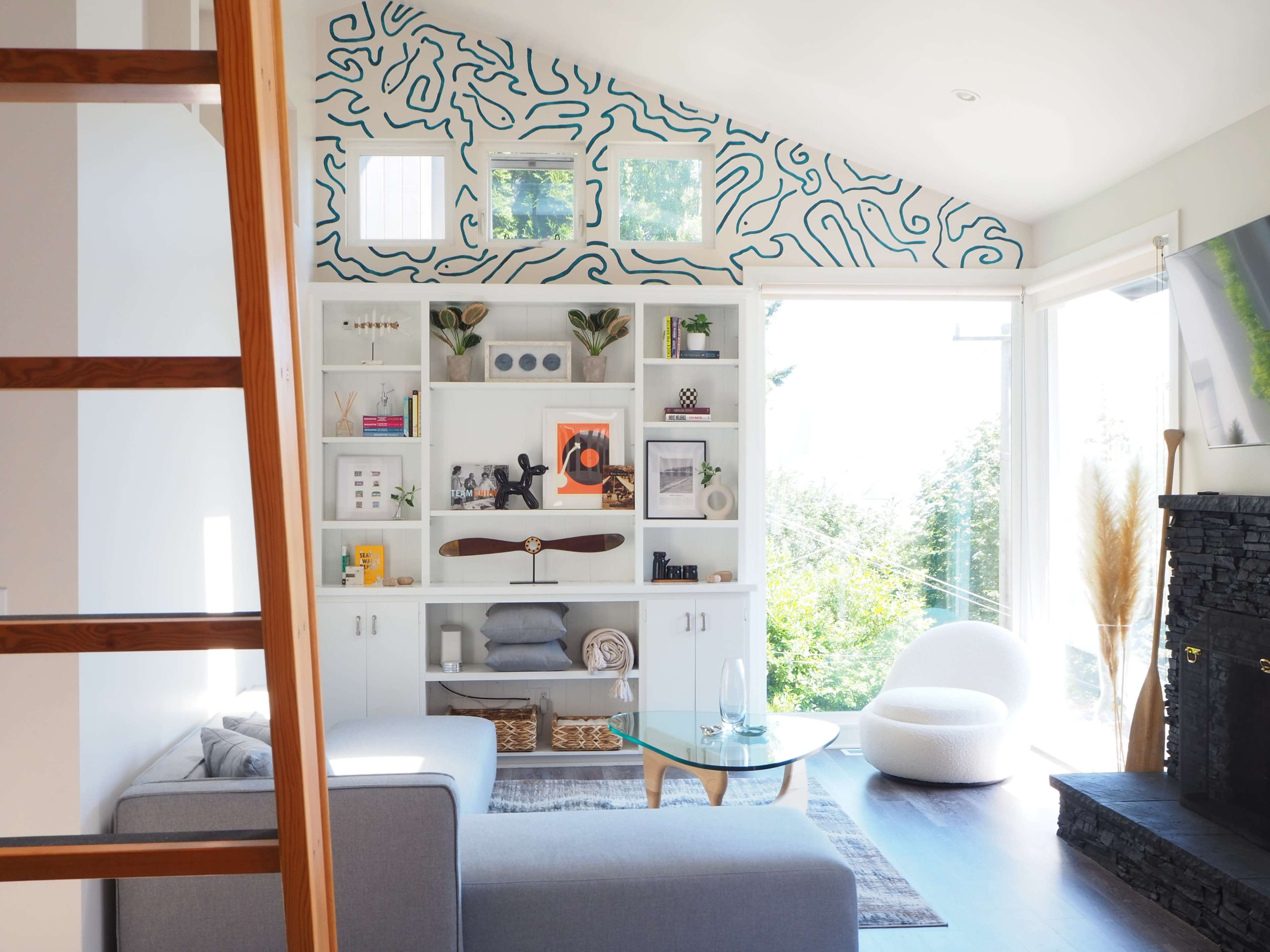 A modern living room with a sectional sofa, a glass coffee table, and a wall featuring a decorative blue line design, alongside a bookshelf filled with decorative items and plants.