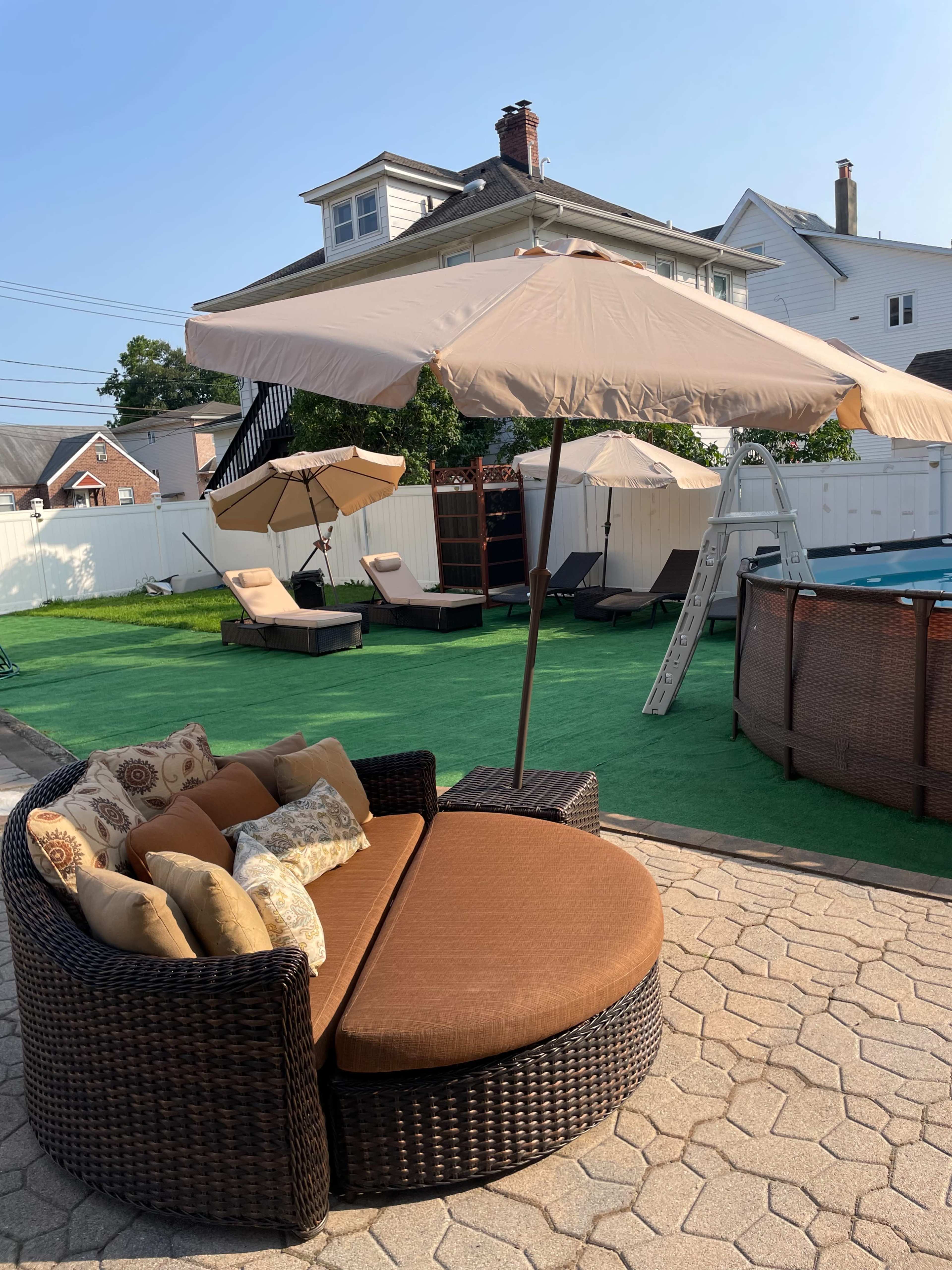 The image shows a patio area featuring a round, wicker outdoor sofa with cushions and an umbrella, surrounded by lounge chairs and a pool.