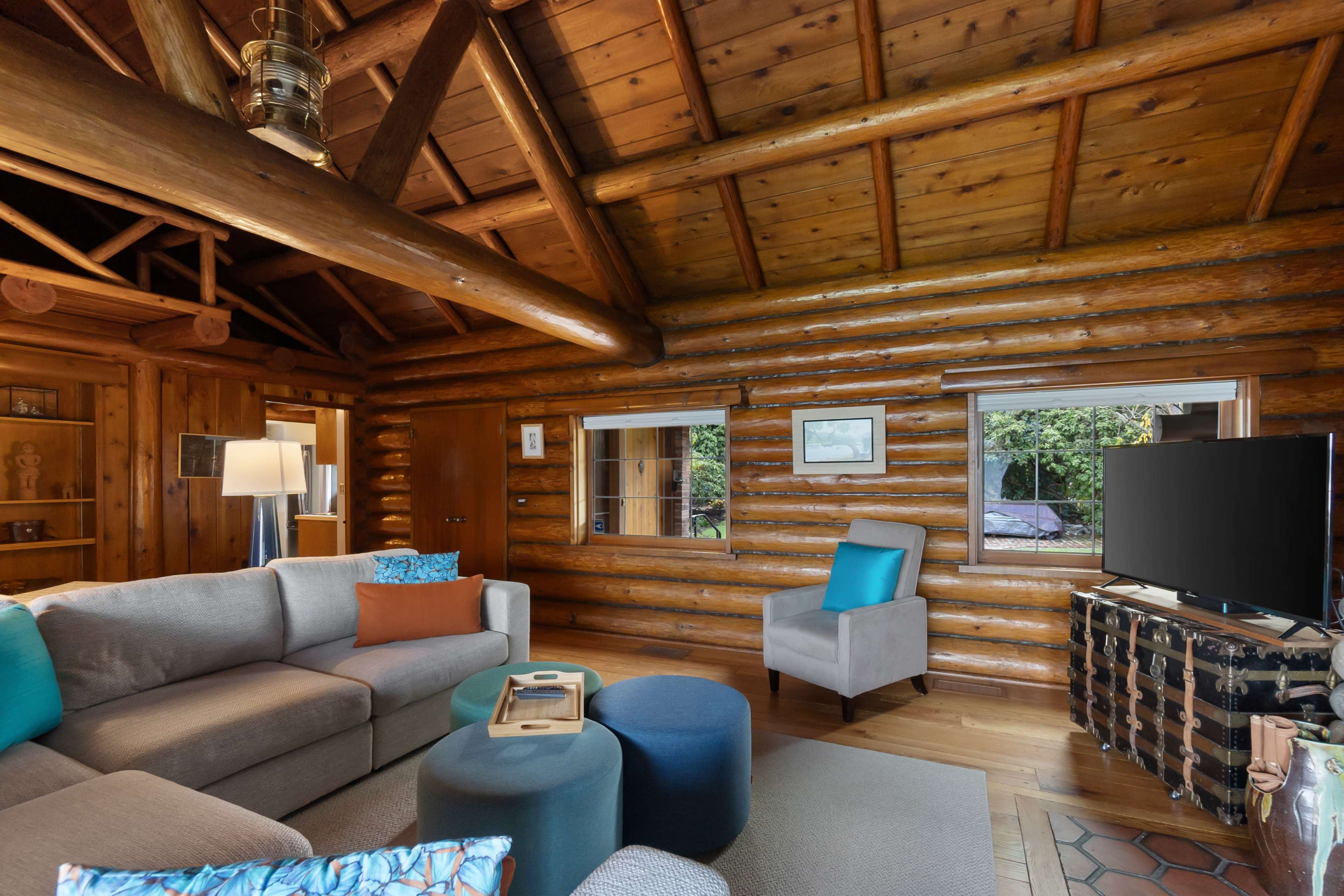 The image shows a cozy living room in a log cabin, featuring a sectional sofa, two ottomans, a television, and wooden walls with large windows revealing greenery outside.