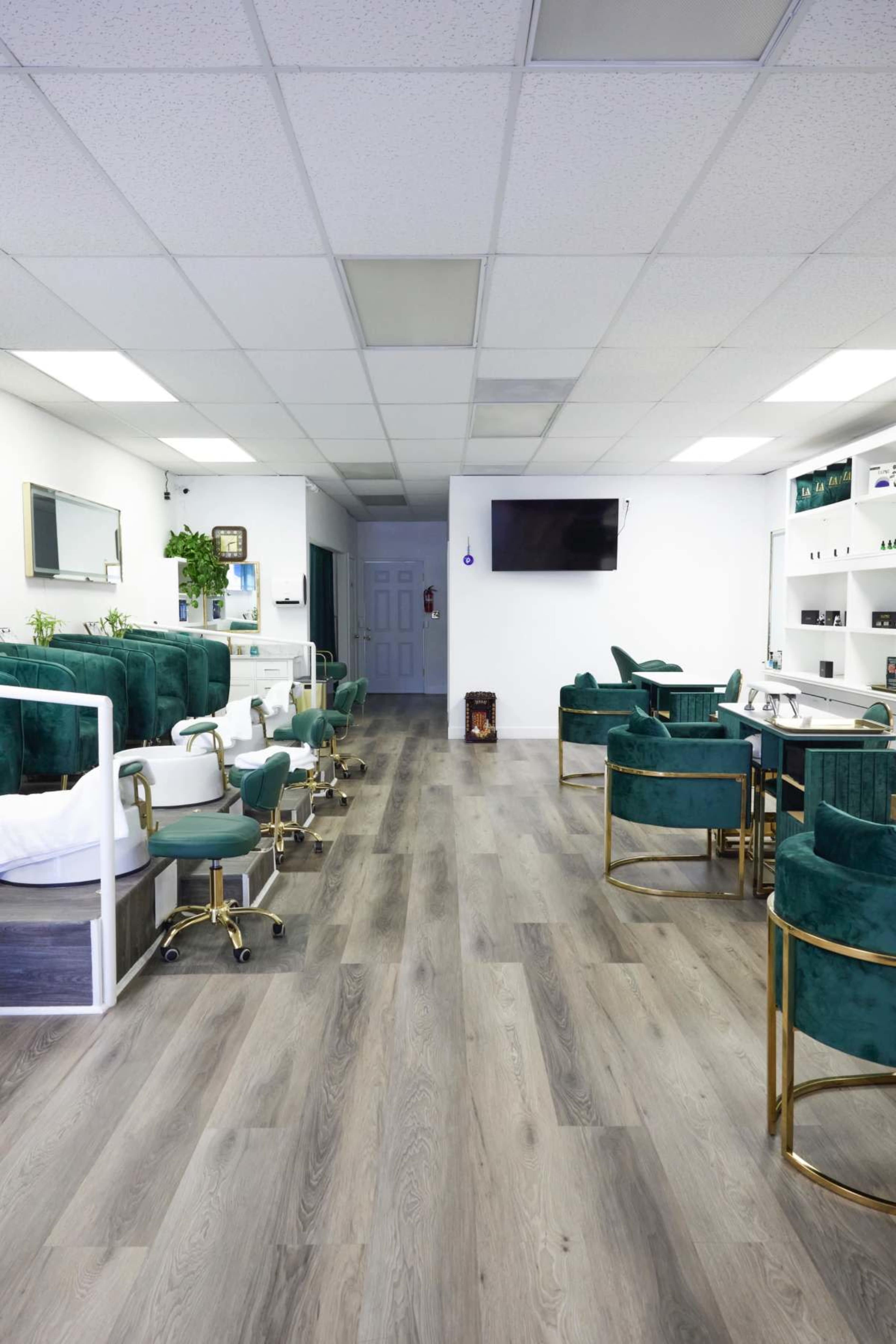 The image shows a modern nail salon featuring green velvet chairs, a series of manicure stations, and a clean, open layout with wooden flooring.
