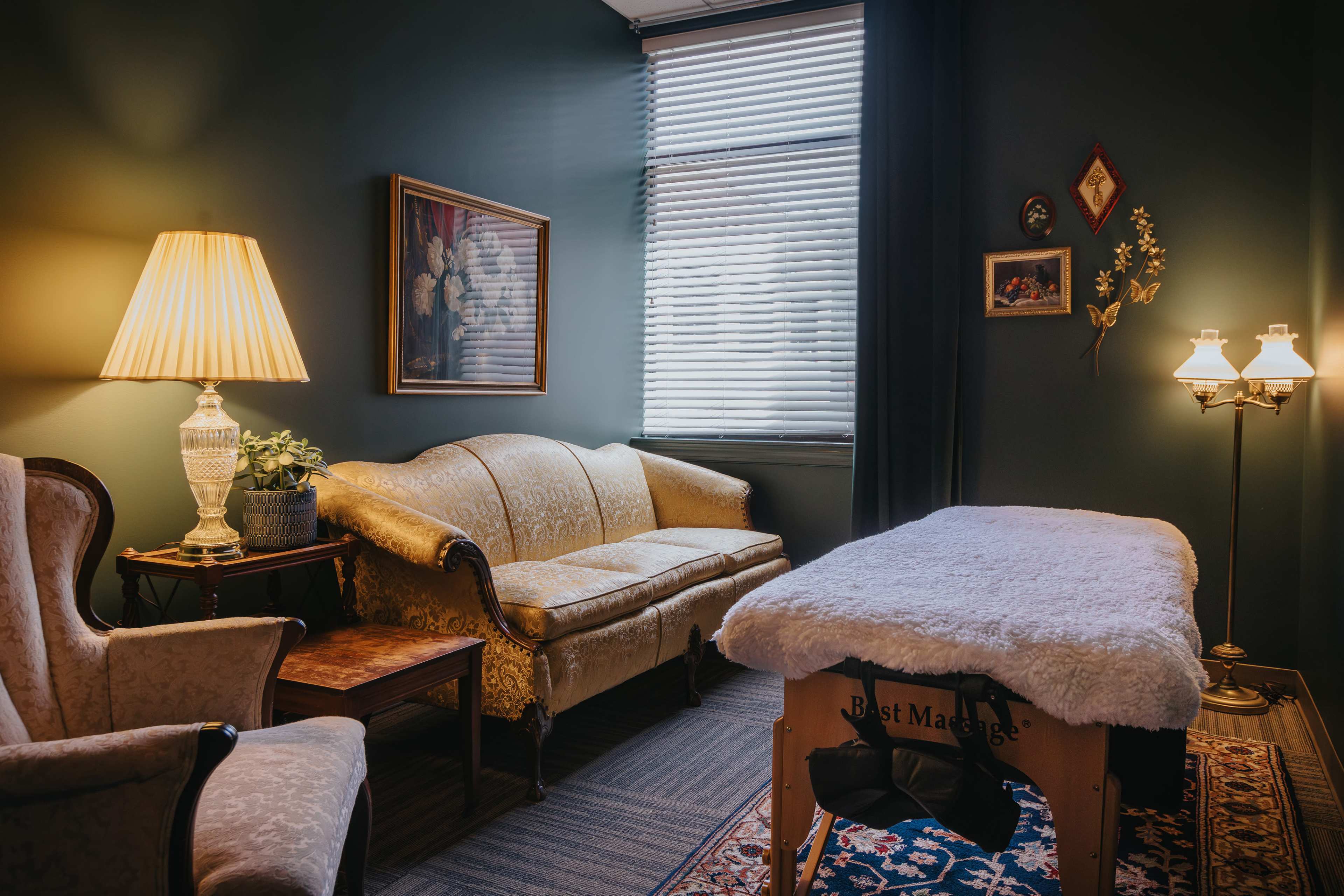 A cozy massage therapy room with a sofa, an armchair, and a massage table draped with a soft covering.