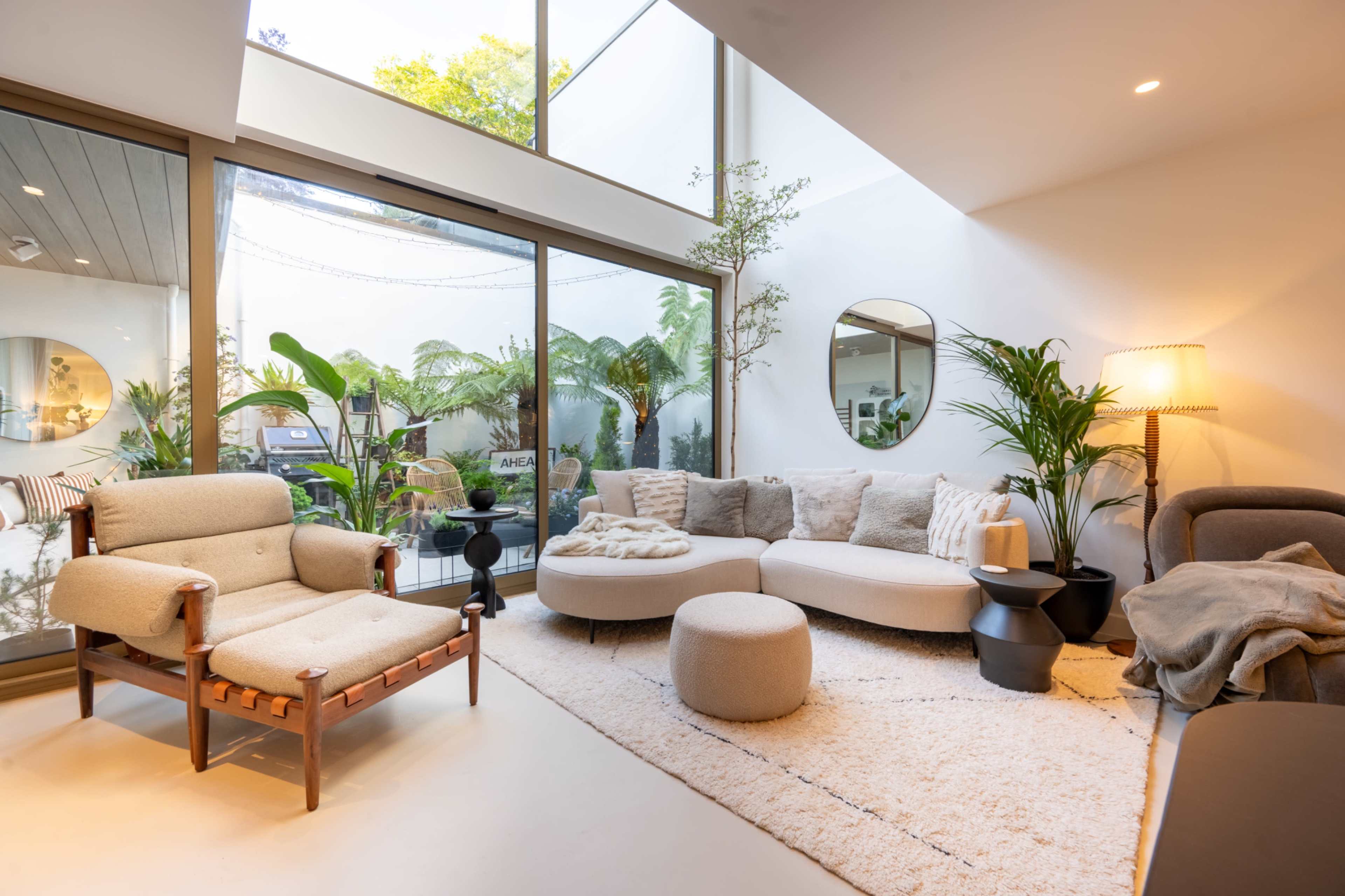 The image shows a modern living room with large glass windows, a curved sofa, a lounge chair, and various indoor plants.