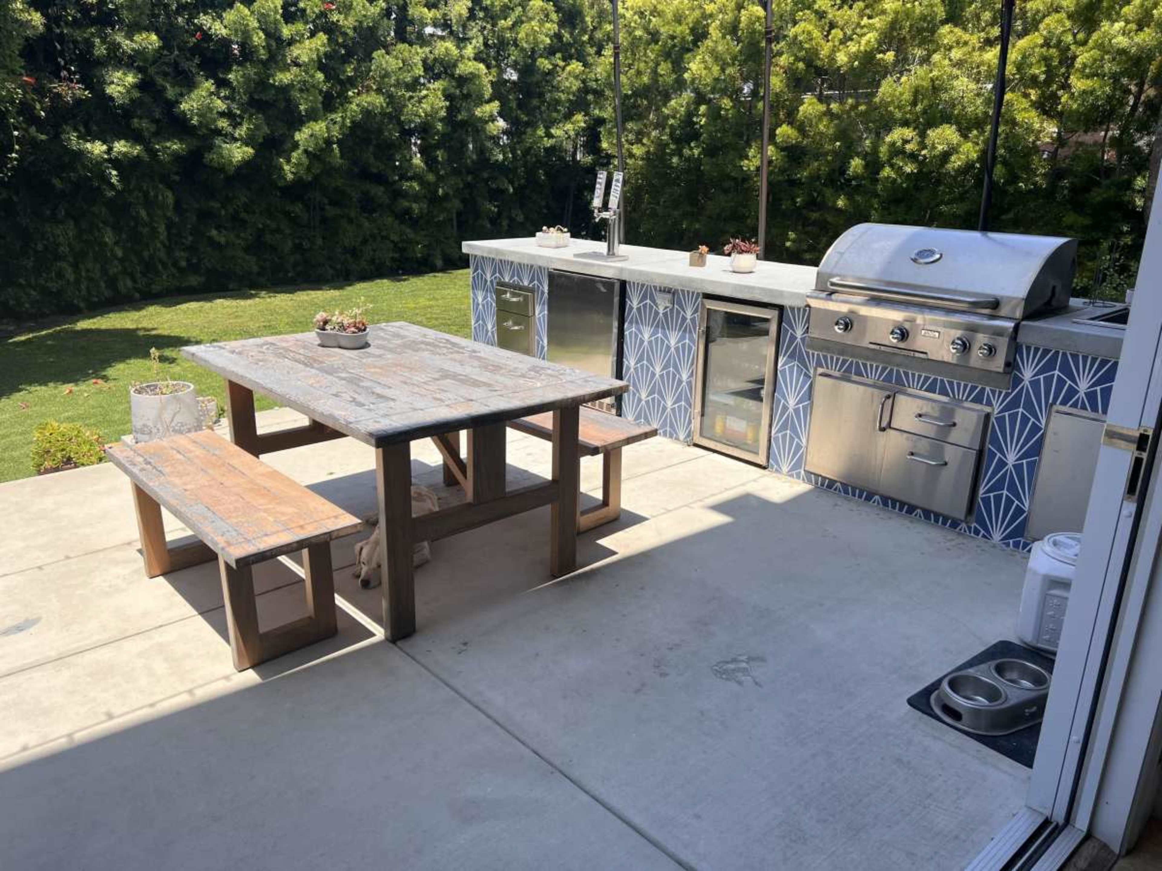 The image depicts an outdoor kitchen area with stainless steel appliances and a wooden dining table with benches on a concrete patio.
