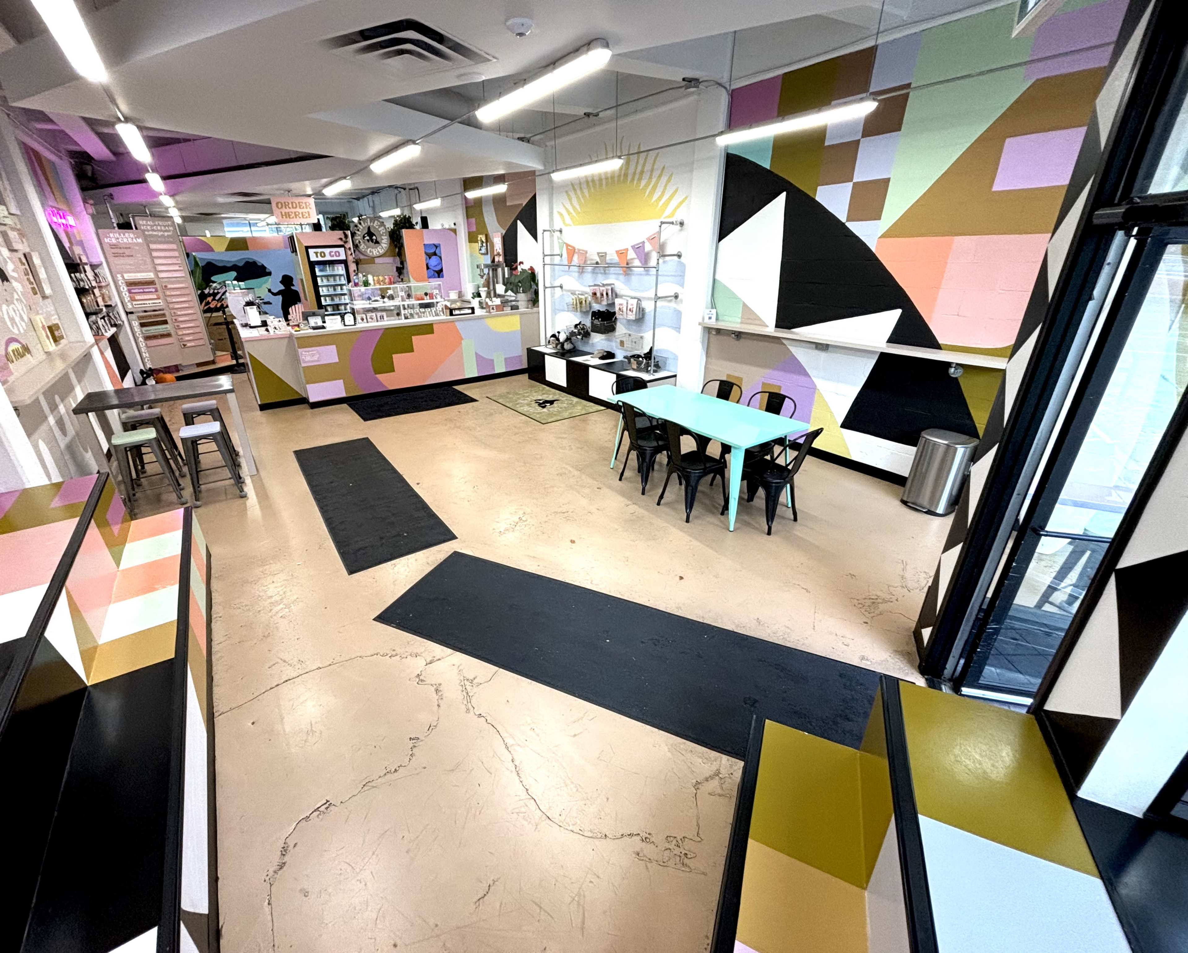 The image shows a modern café interior with colorful geometric wall murals, a service counter, and a mix of seating options.
