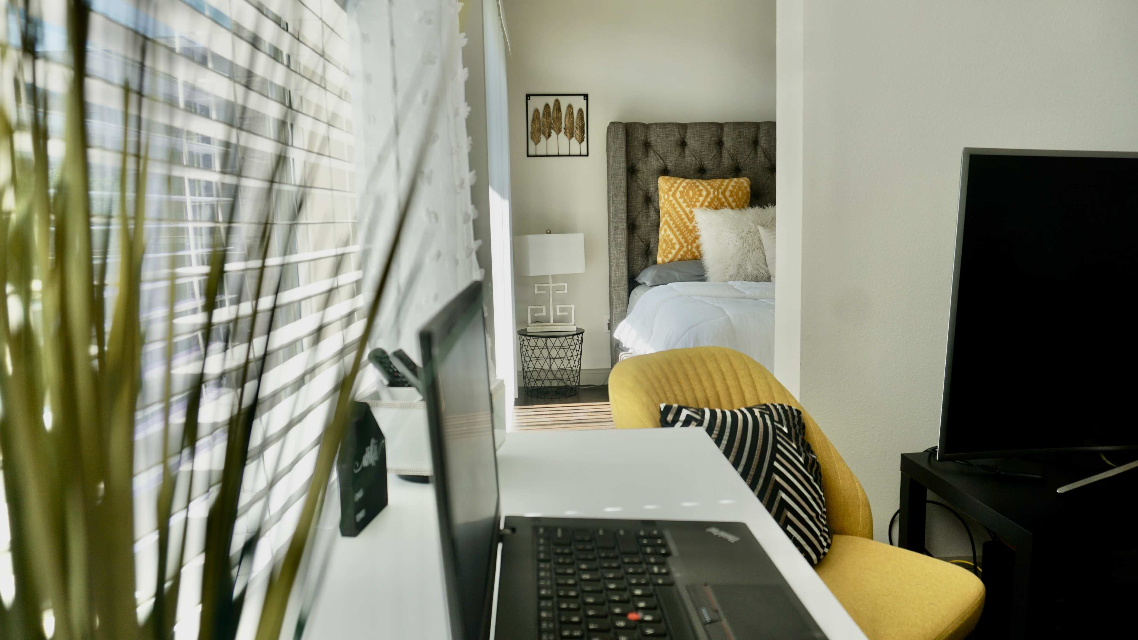 A modern workspace features a laptop and chair in the foreground, with a neatly made bed visible in the background through a doorway.
