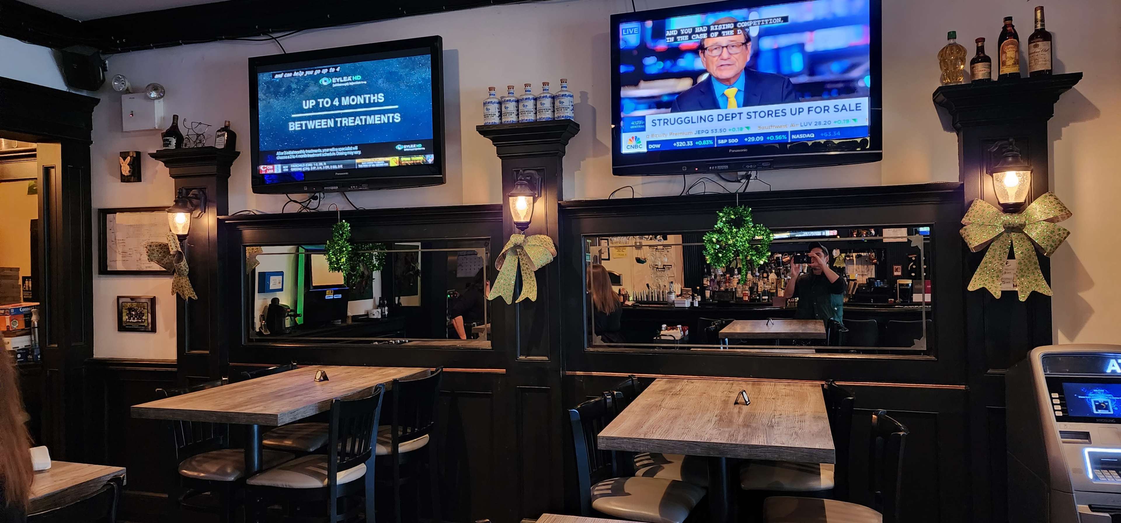 A restaurant interior featuring two mounted televisions displaying news, wooden tables, and festive green decorations on the mirrors.