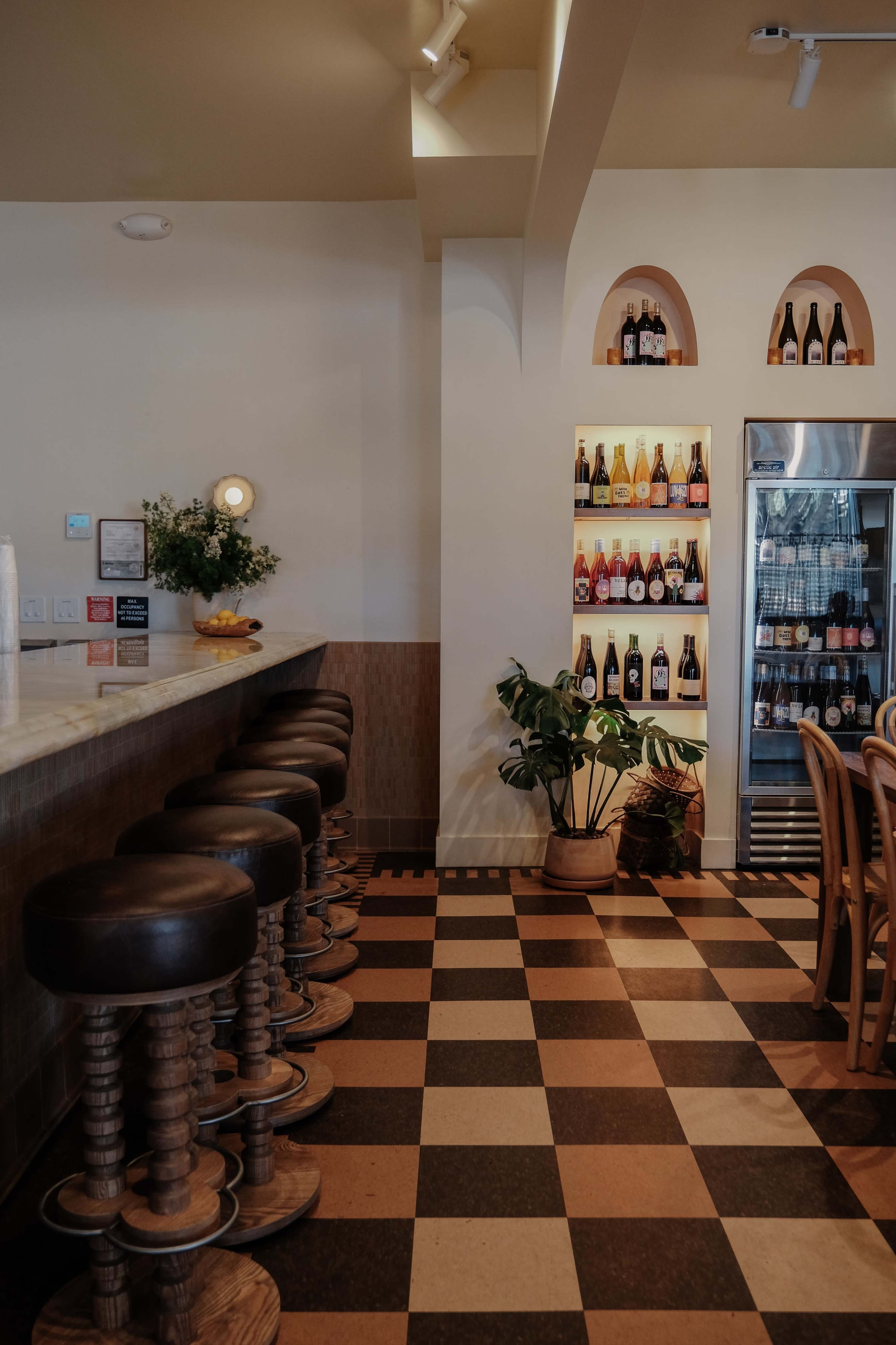 A bar area with a long countertop, wooden stools, a plant, and a wall displaying various bottles in a refrigerator and on shelves.