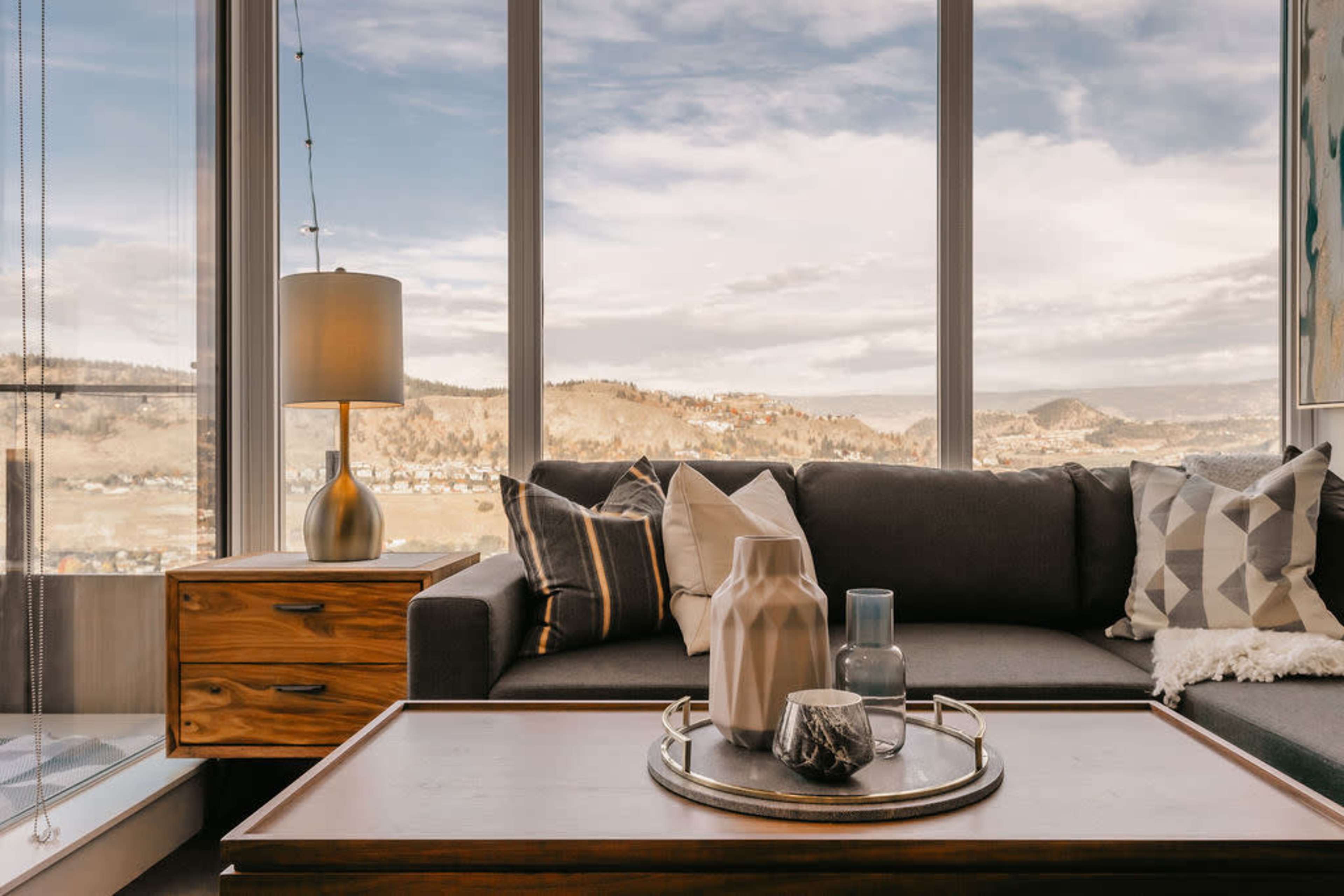 The interior features a modern living room with a gray sofa, a wooden side table, and large windows showing a view of mountains and a cloudy sky.