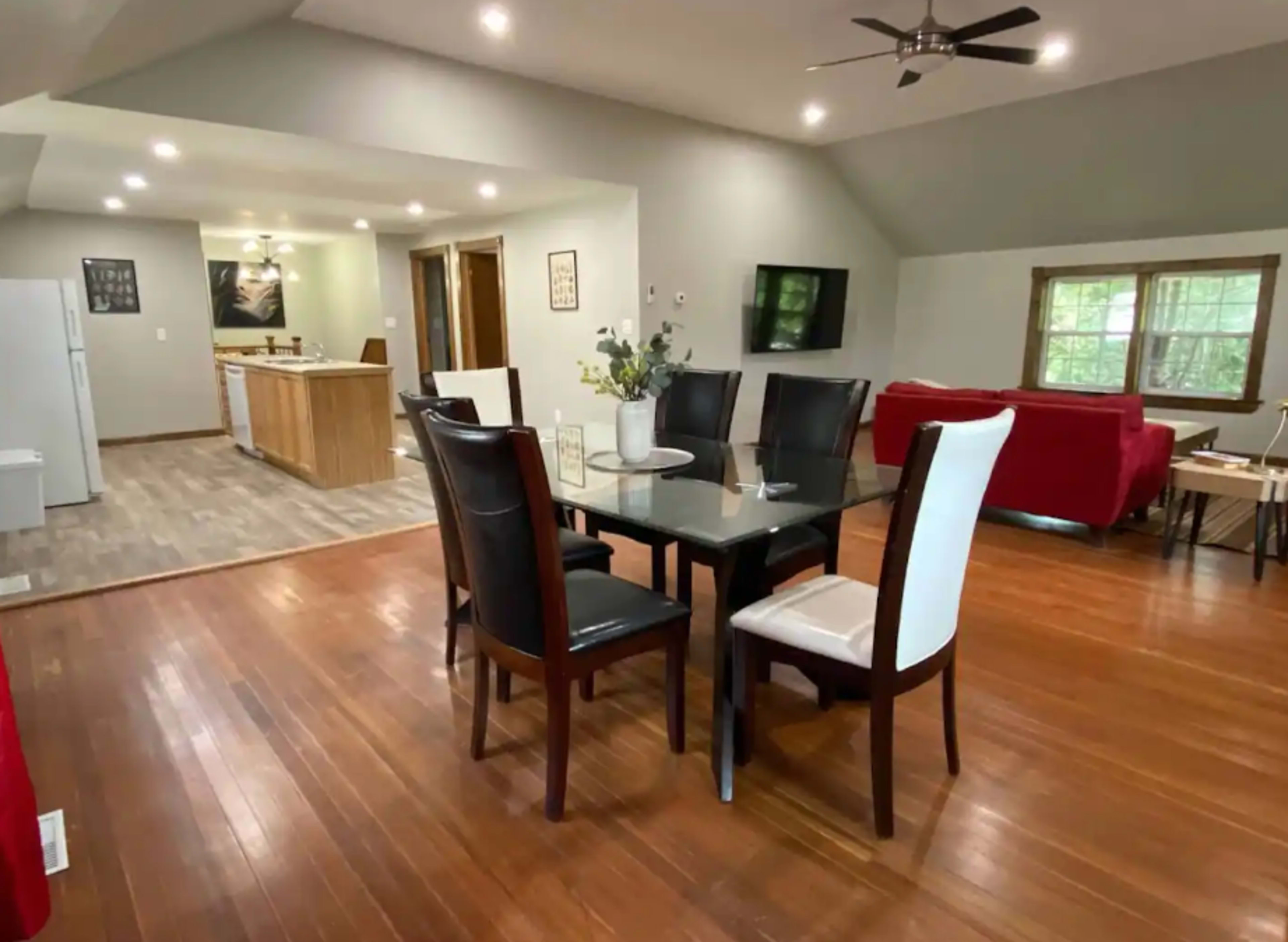 The image shows a spacious dining area with a glass-top table surrounded by black and white chairs, leading into a modern kitchen and a living room with a red couch.