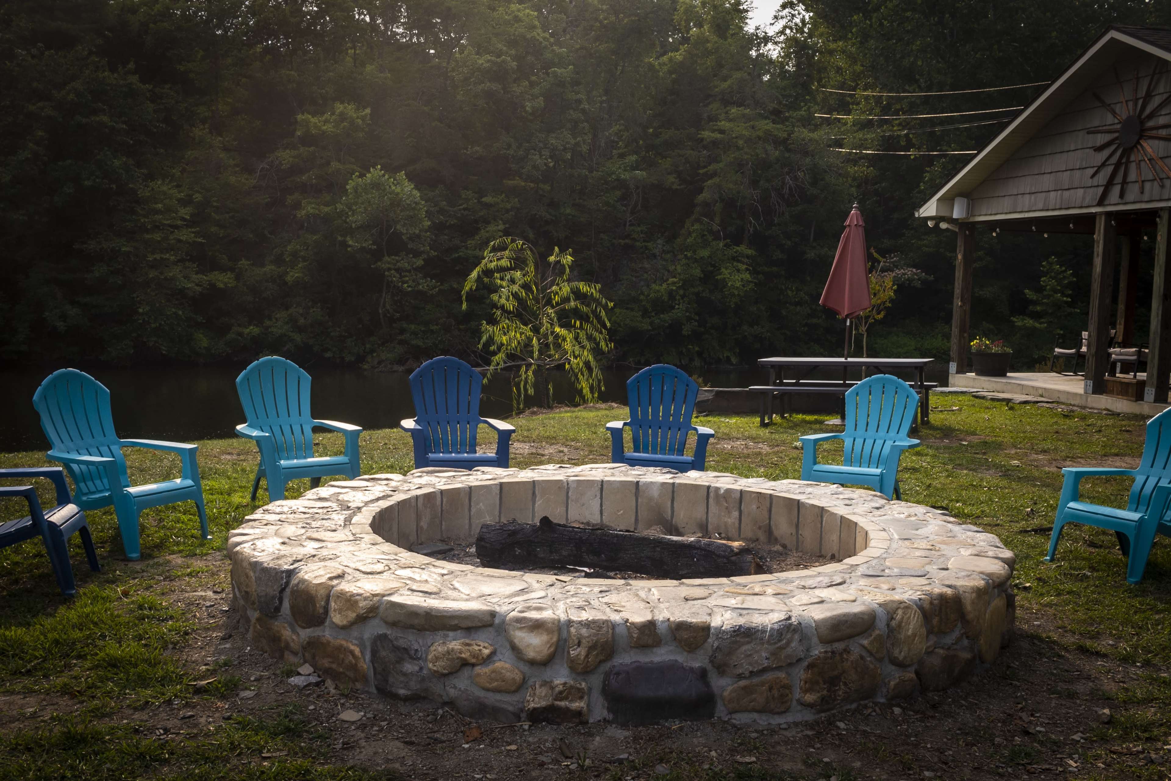 A circular stone fire pit is surrounded by blue plastic chairs near a serene body of water and a cabin in a wooded area.